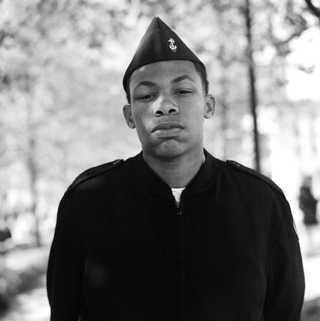 A young man in a dark military-style uniform and cap stands outdoors, looking directly at the camera. Trees and sunlight create a softly blurred background.
