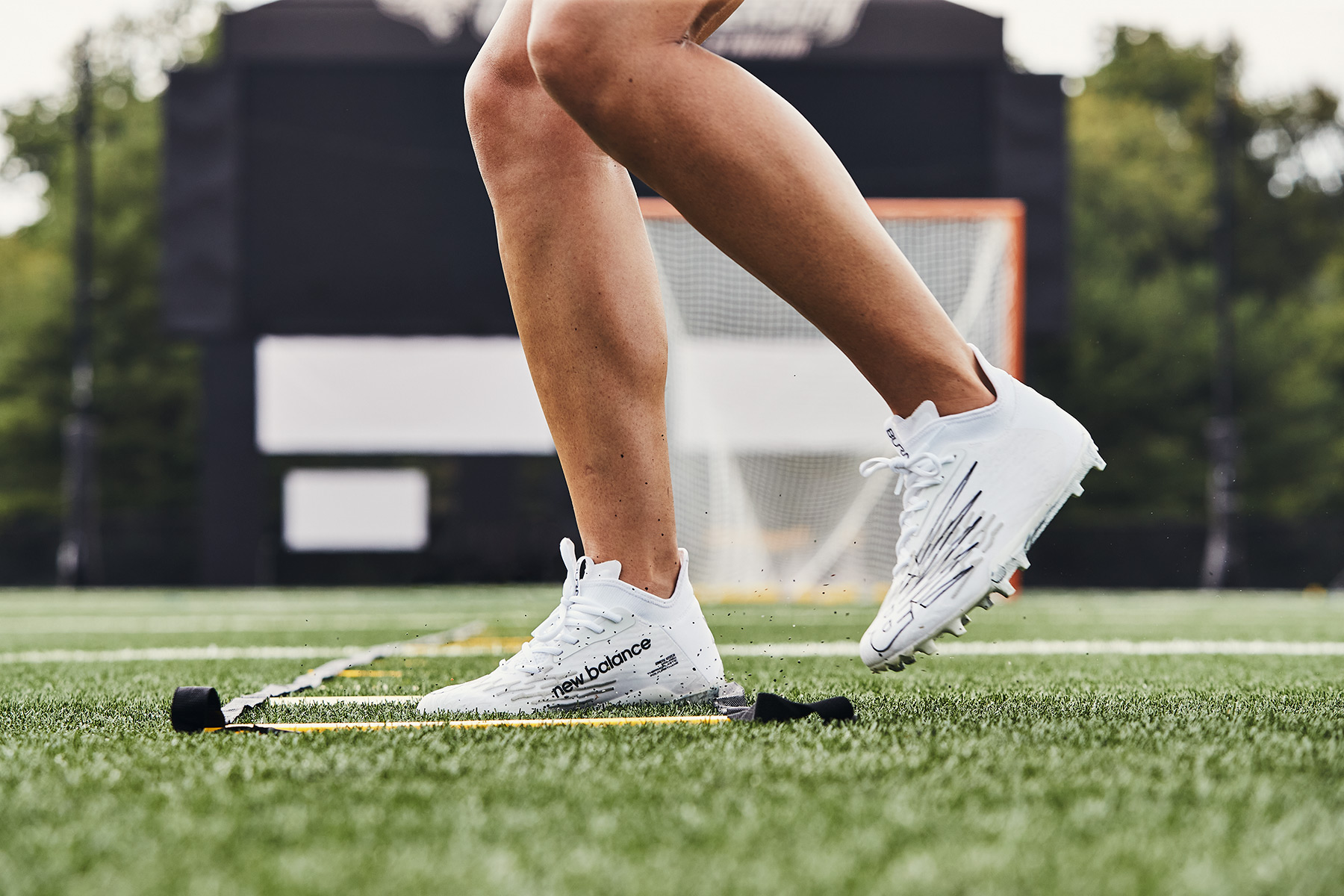 A person wearing white athletic shoes with New Balance branding performs agility ladder drills on a green turf field, brought to life with CGI graphics for product marketing; sports equipment and trees blur in the background.