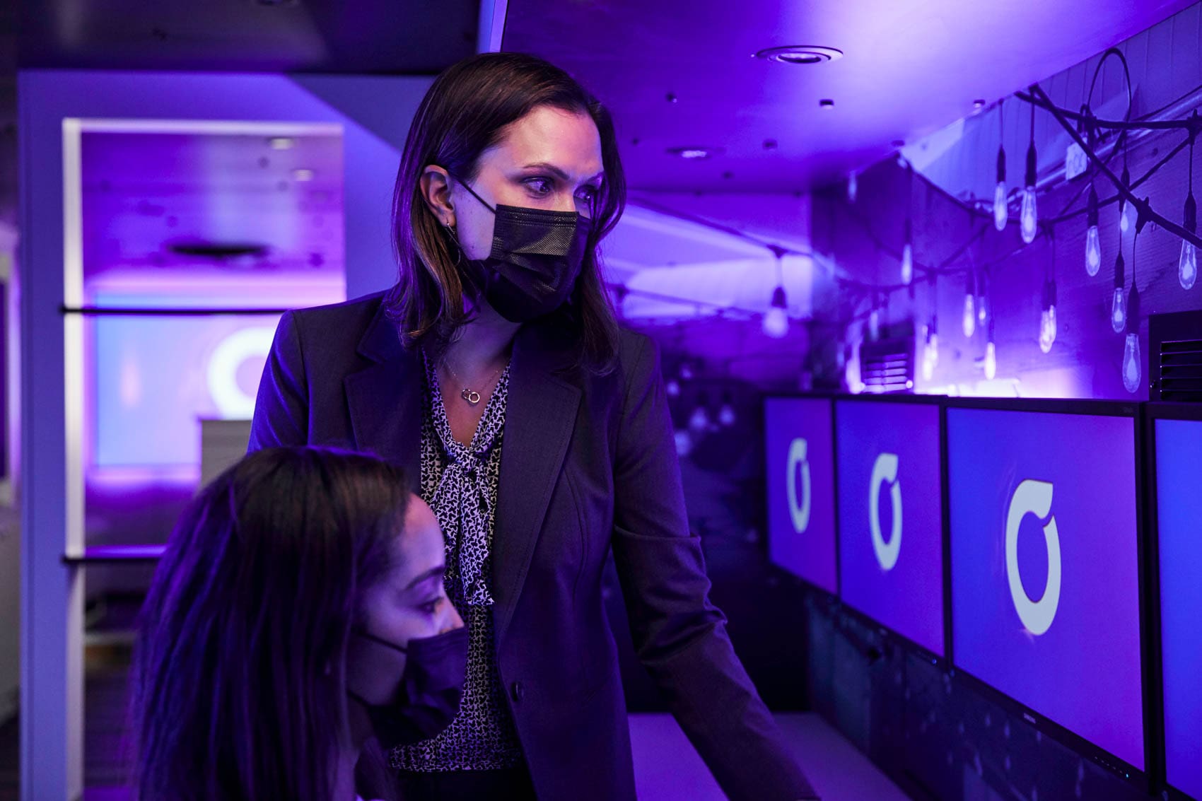 Two women wearing masks work in a room with purple lighting, looking at multiple computer monitors displaying a circular logo. One woman is standing, the other is seated, both focused on the screens.