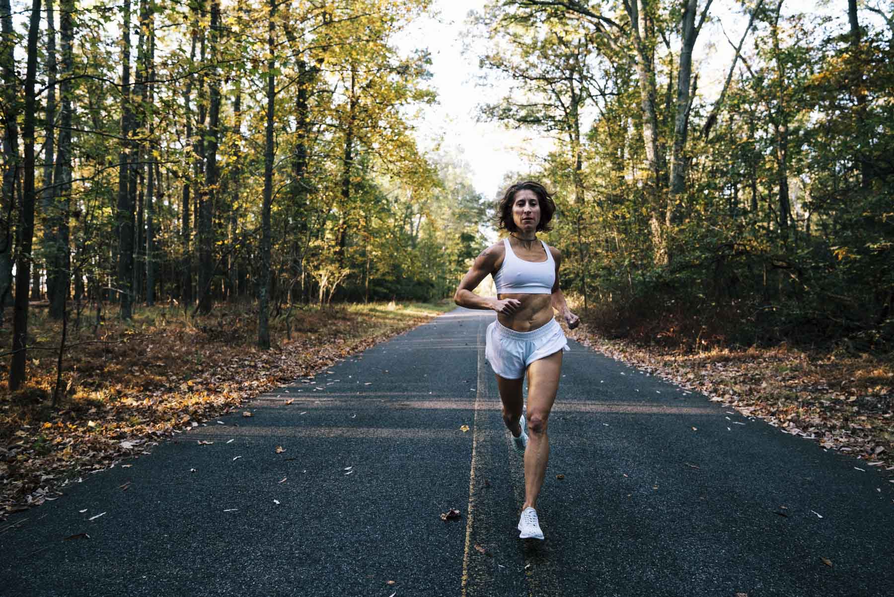 A woman in athletic wear runs on a paved path through a forest with autumn trees, listening to music with wired earphones. The sun shines through the trees, creating a peaceful outdoor setting.