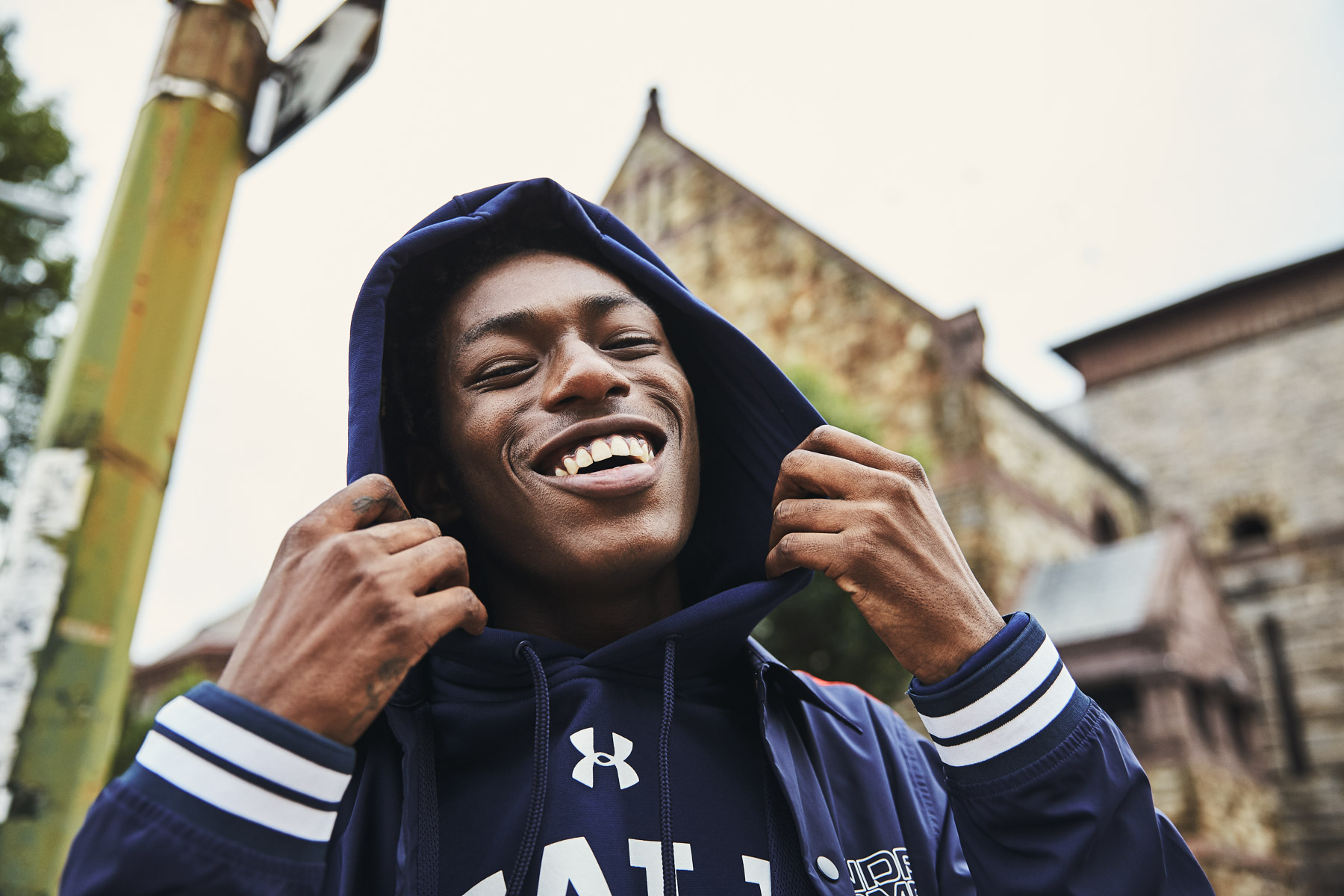 A young man wearing a navy blue hoodie and jacket smiles joyfully, holding the hood over his head. He stands outside near a building with stone walls and greenery in the background.