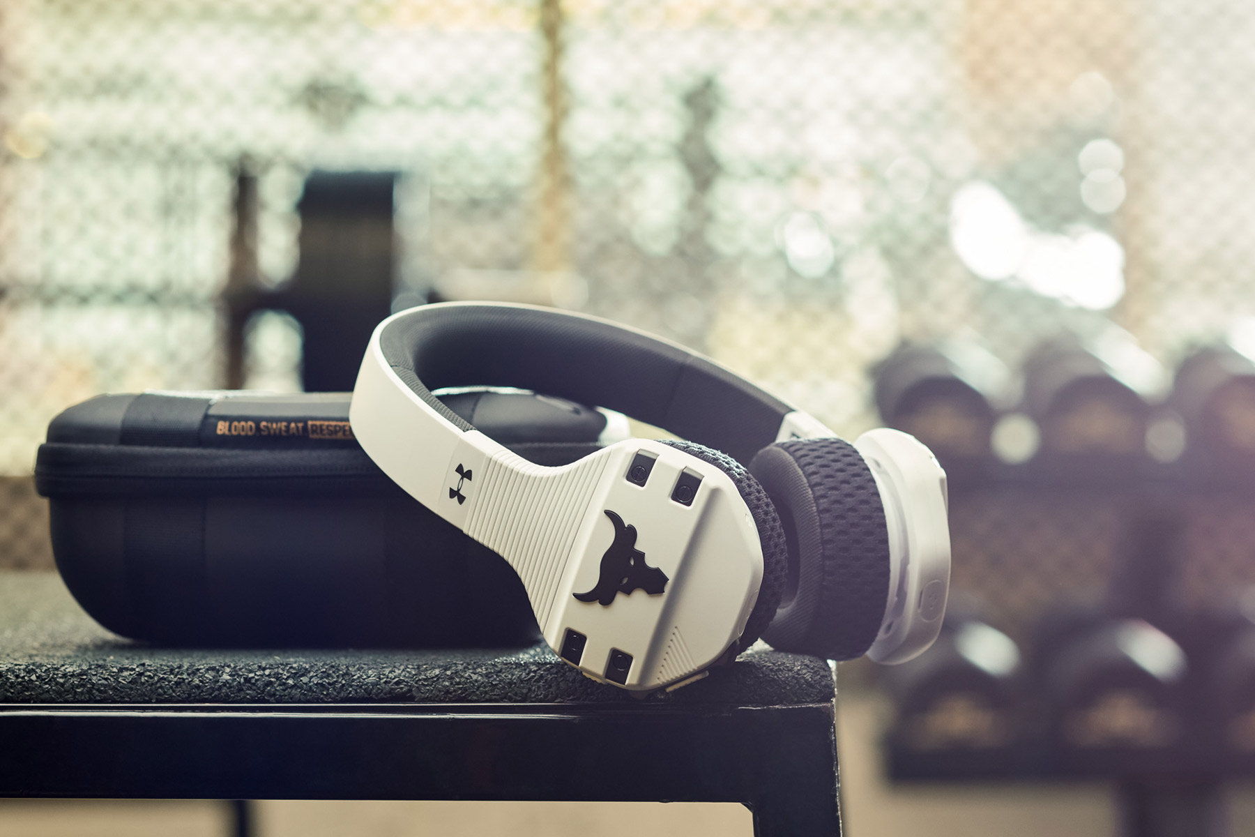 White and black wireless headphones with a bull logo rest on a hard case atop gym equipment, with blurred dumbbells and a mesh fence in the background.