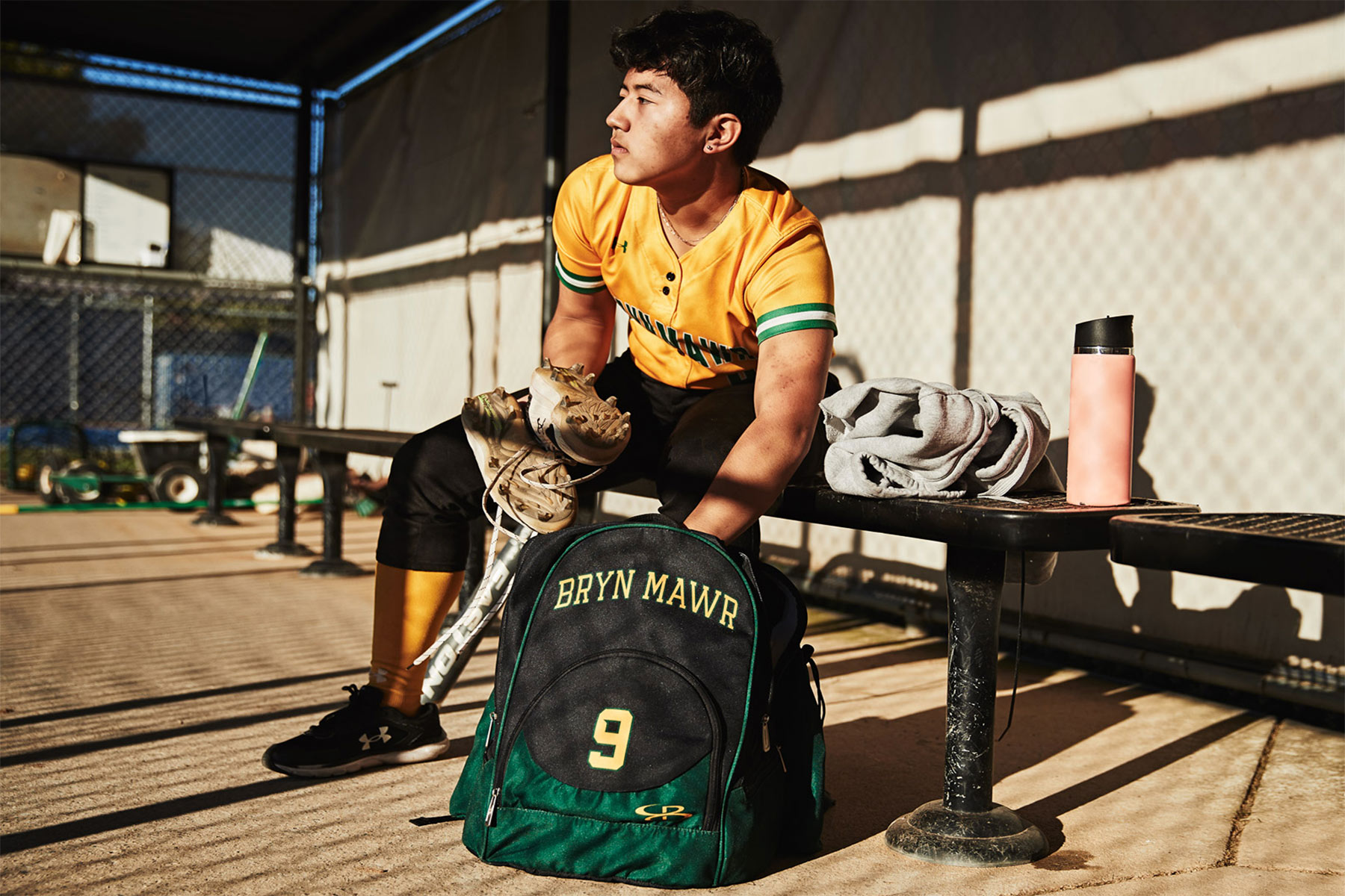 A baseball player in a yellow uniform sits on a bench in the dugout, holding a glove and gazing into the distance. Captured by a Washington DC photographer, his green Bryn Mawr backpack with number 9 and pink water bottle complete this advertising sports scene.