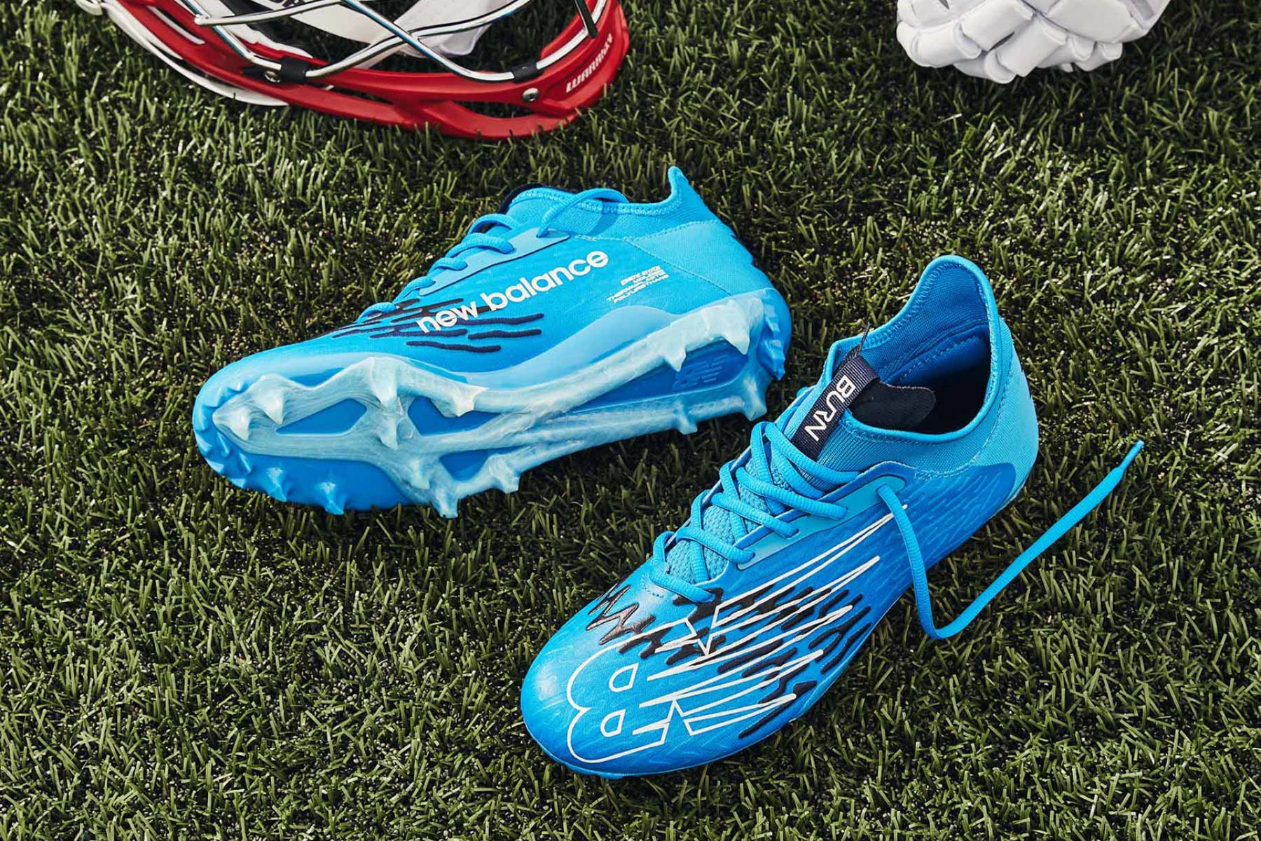 A pair of new balance cleats rest in the grass on a field with other lacrosse equipment.