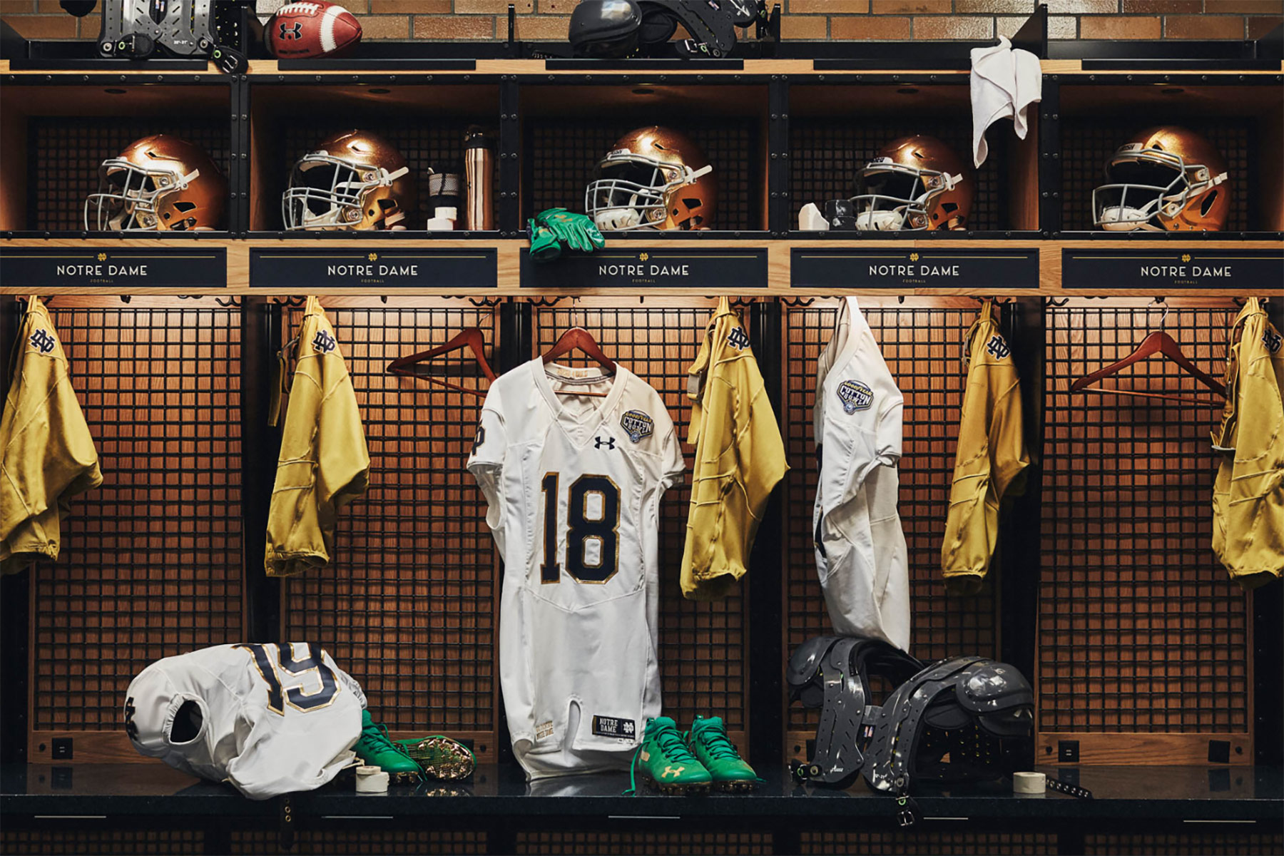 Notre Dame football locker room with three open lockers displaying gold helmets, white and gold jerseys, shoulder pads, green cleats, and other gear, with a white jersey number 18 hanging in the center.
