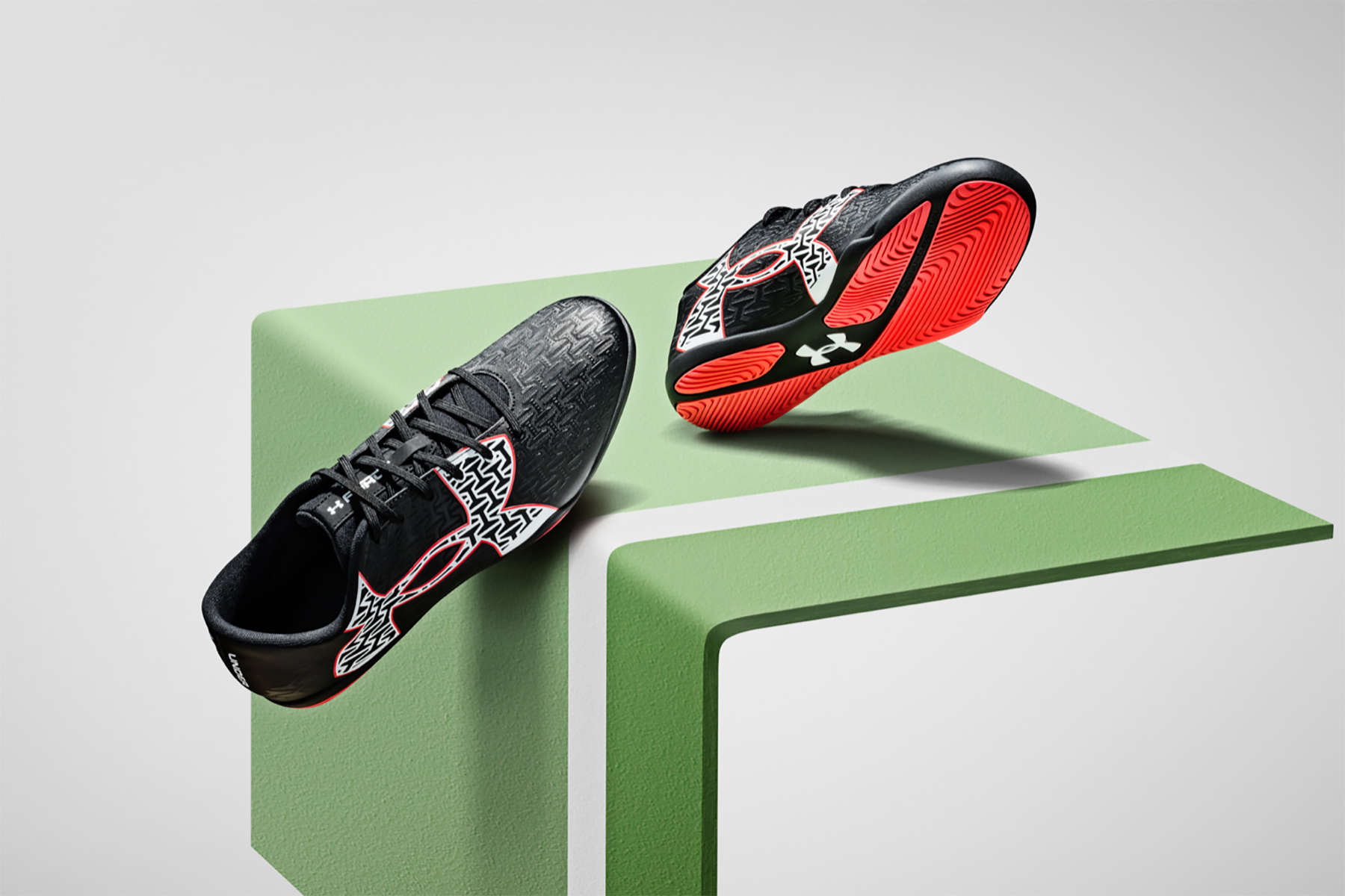 Two black athletic shoes with red and white abstract patterns are displayed on a green and white geometric platform, with one shoe upright and the other showing its red sole. The background is plain and light-colored.