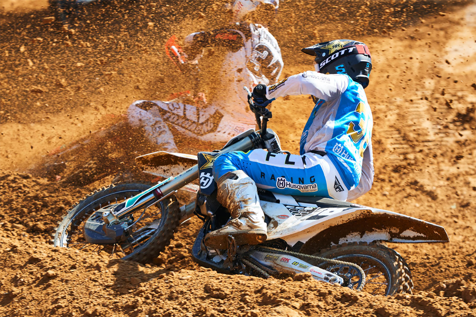 Two motocross riders race through a muddy dirt track, kicking up a spray of mud. The rider in front wears blue and white gear with a black helmet, leaning into a turn, with another racer partially visible behind.