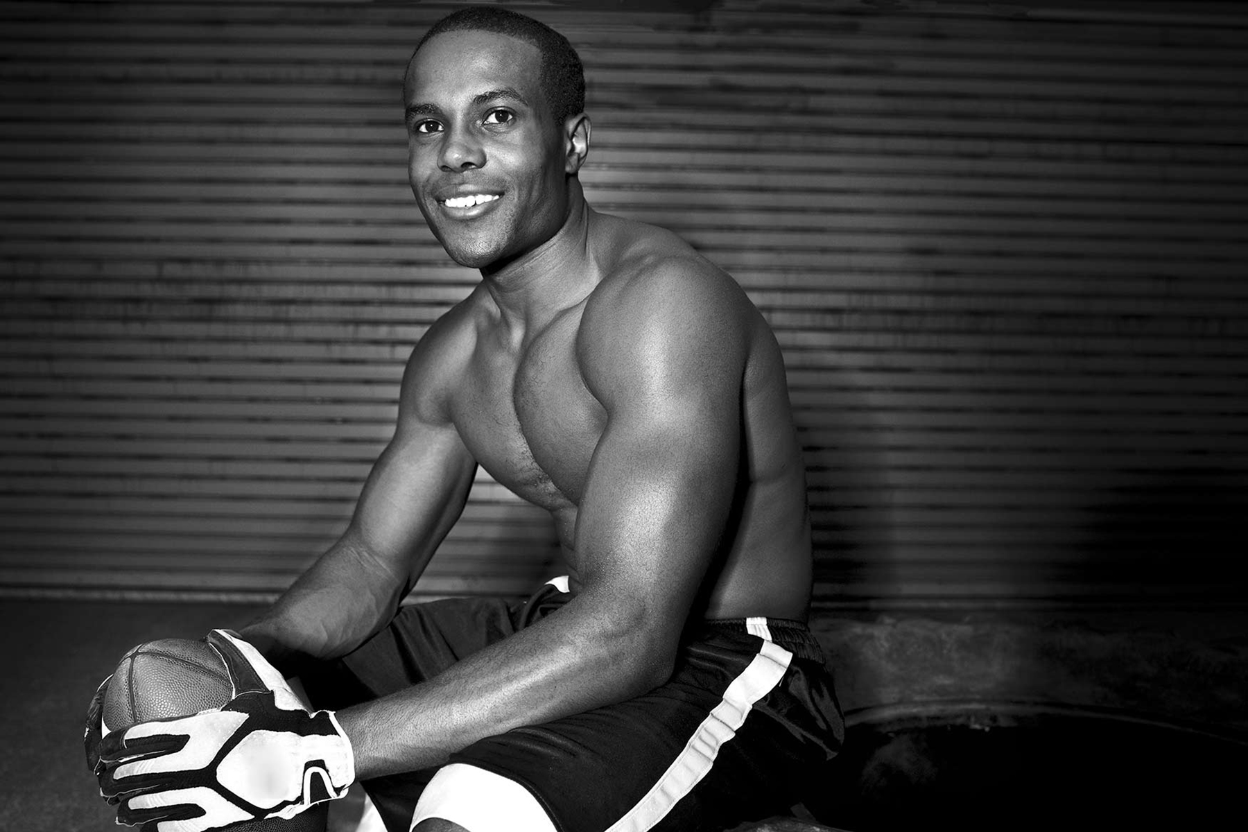 A shirtless man wearing sports gloves and shorts sits while holding a football, smiling at the camera. Captured by a sports advertising photographer, the black and white image features a corrugated metal wall in the background.