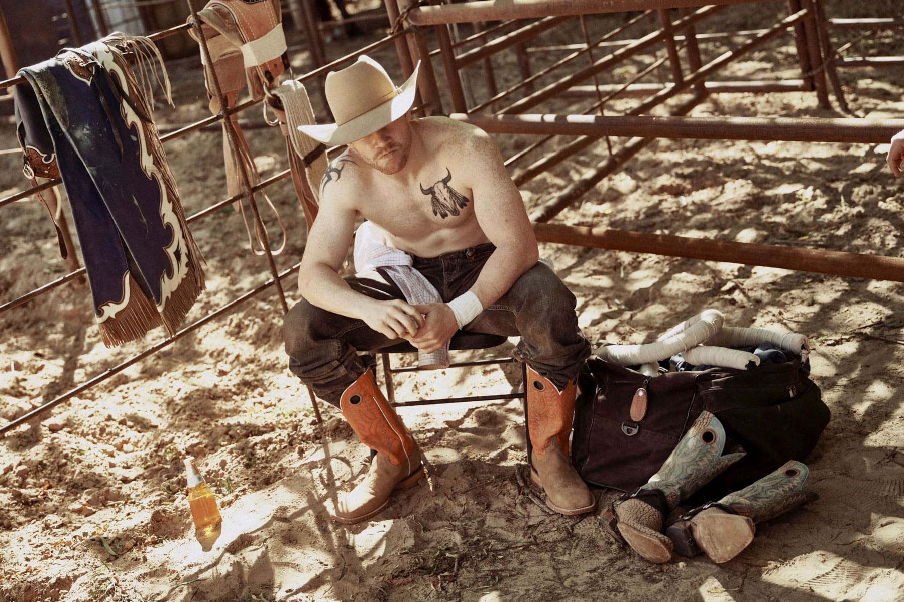 A shirtless cowboy, inspired by Travis Gillett’s ranching spirit, sits on a chair in a sandy rodeo pen, looking down. Gear, boots, and a bag lay nearby, with a bottle and western clothes hanging on the fence.