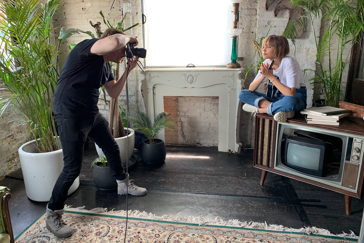 A photographer takes a picture of celebrity, Grace Vanderwaal, sitting cross-legged on a vintage TV set in a bright, plant-filled room with a fireplace and decorative items.