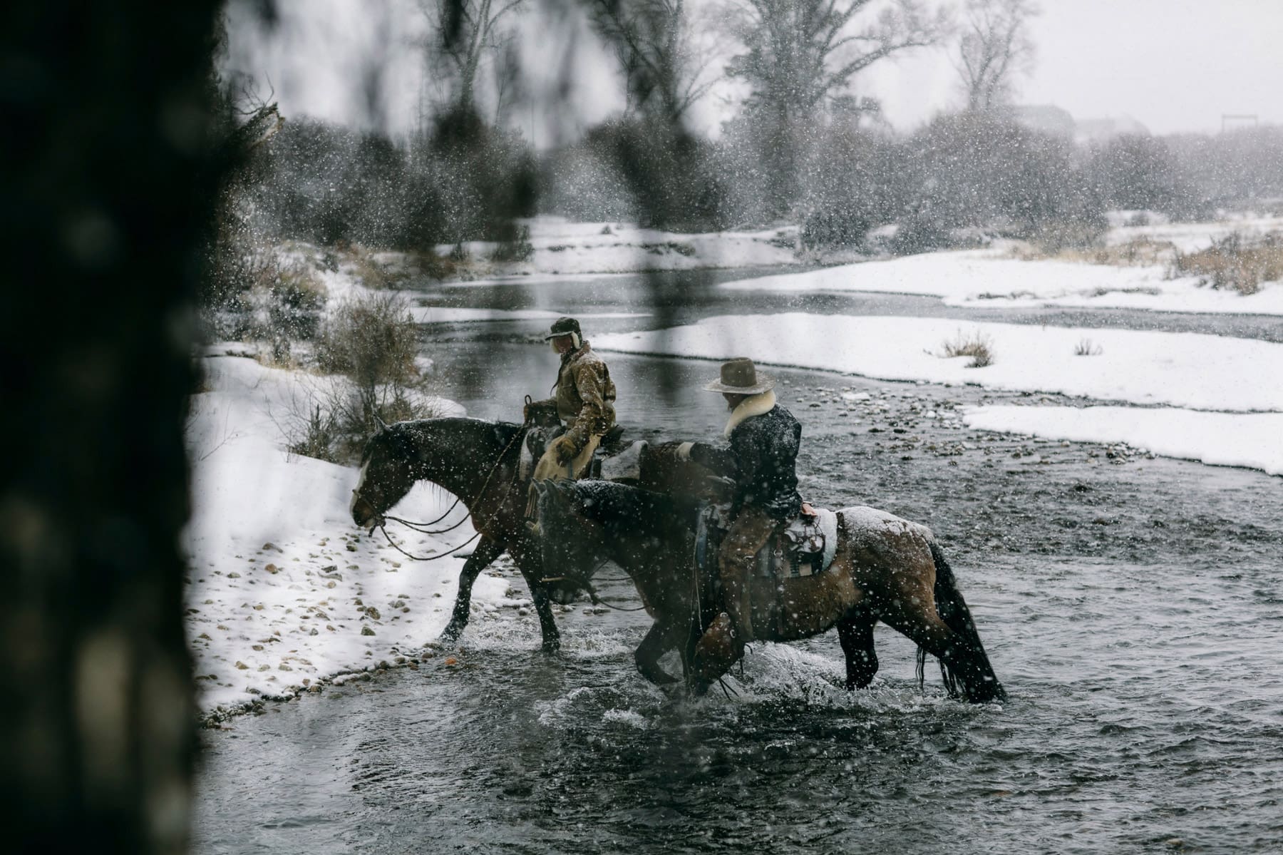 Two men on horses take their horses across a snowy river in Washington.