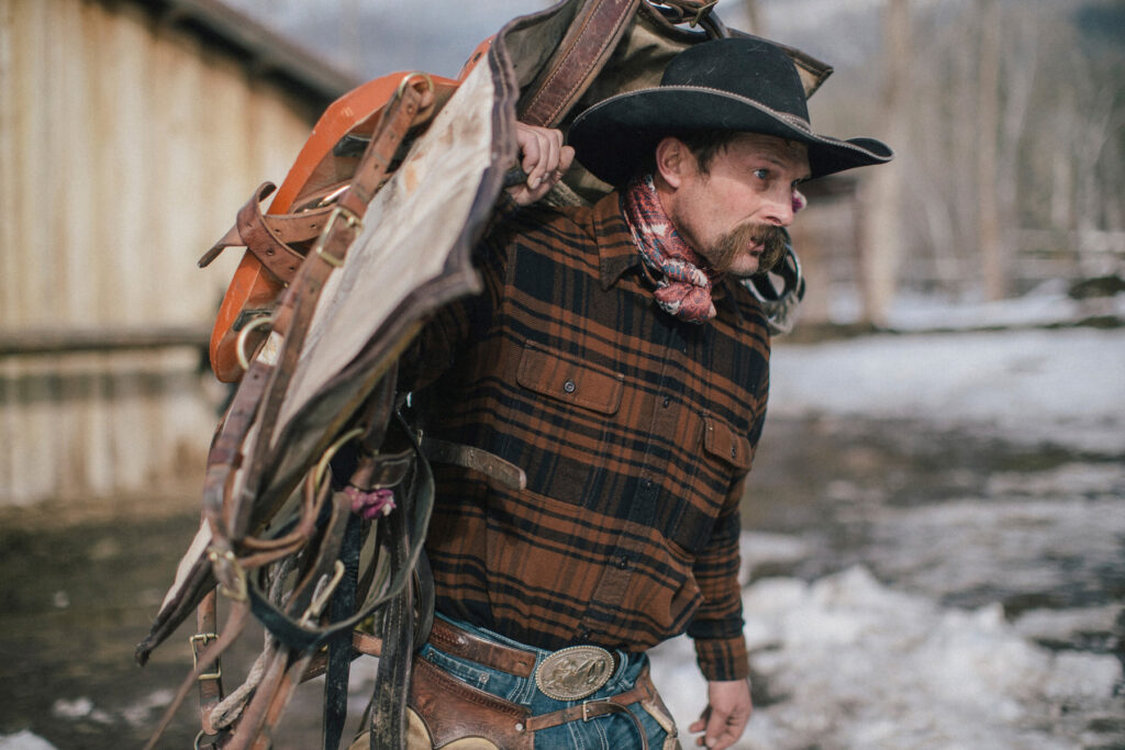 Pictured, a man brings a saddle over to his horse in the snowy landscape of the American West.