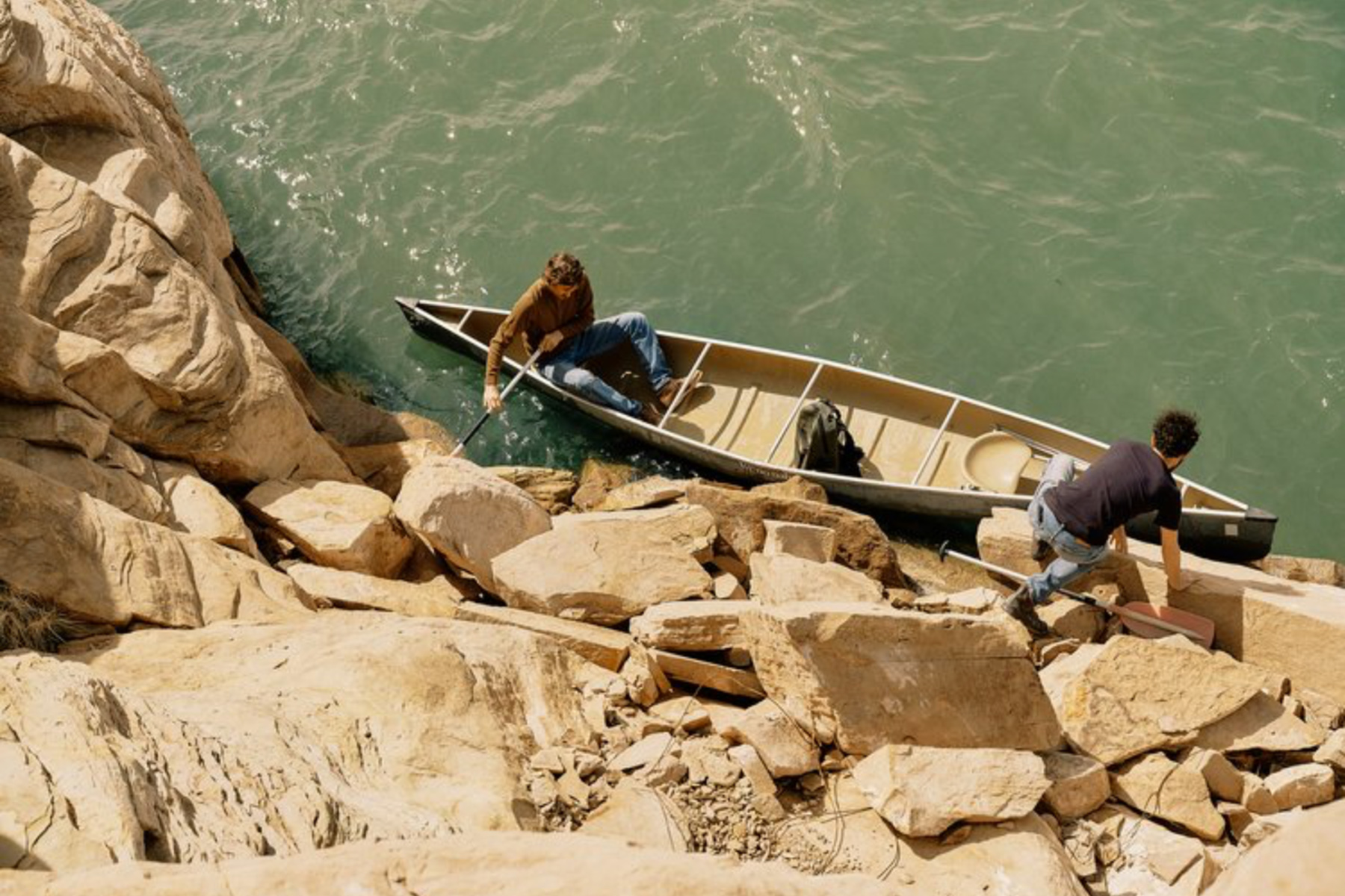 Two people are climbing over large rocks on a riverside; one person is in a canoe holding onto the rocks while the other is stepping onto the rocky shore from the water.