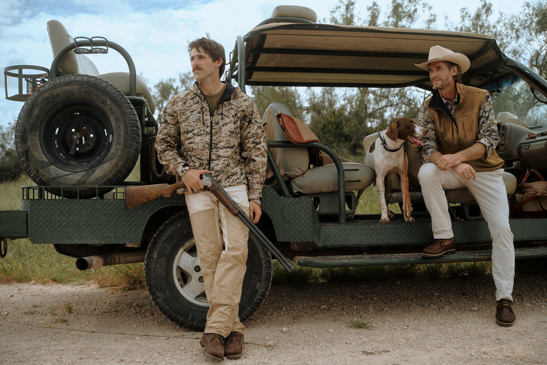 Two men in outdoor attire pose by a green safari vehicle in Texas; one stands with a rifle while the other, accompanied by a dog, sits on the back—captured through the lens of hunting fashion photographer Travis Gillett. Grass and trees fill the background.