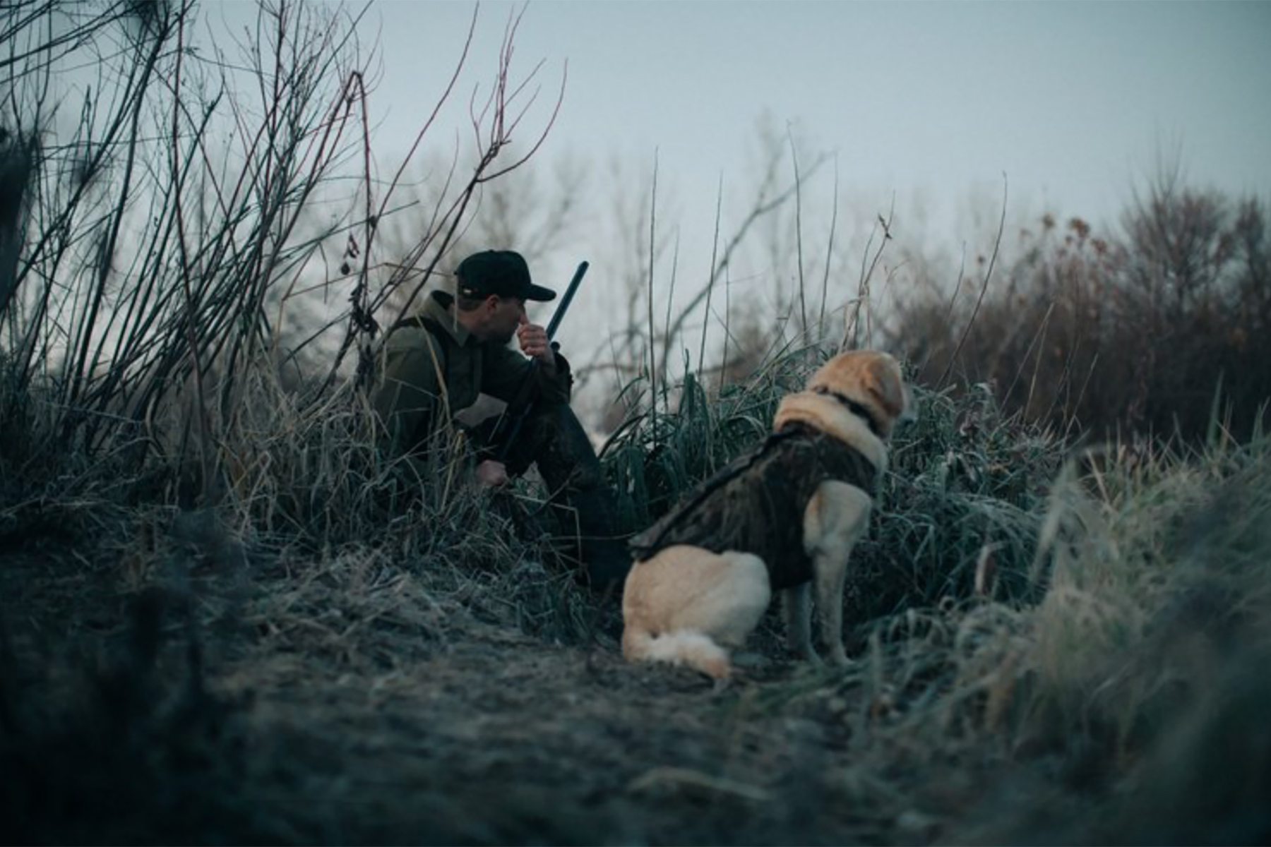 A person in camouflage sits with a rifle among frosty grass, joined by a yellow dog in a vest—both gazing intently ahead. Captured by hunting and fishing photographer Travis Gillett, this cold outdoor moment spans from Seattle to Texas.