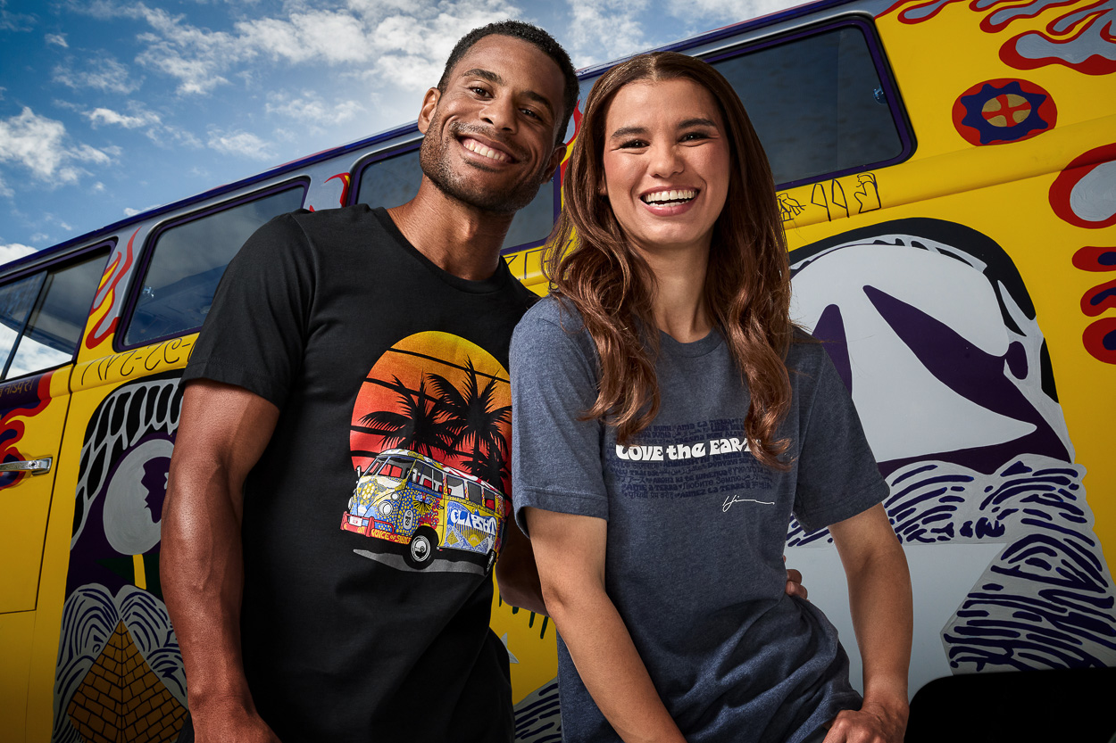 A smiling man and woman stand in front of a colorful, retro-style van with bold, psychedelic designs. Both wear graphic T-shirts; the man’s has a sunset and palm trees, the woman’s says “LOVE THE EARTH.”.