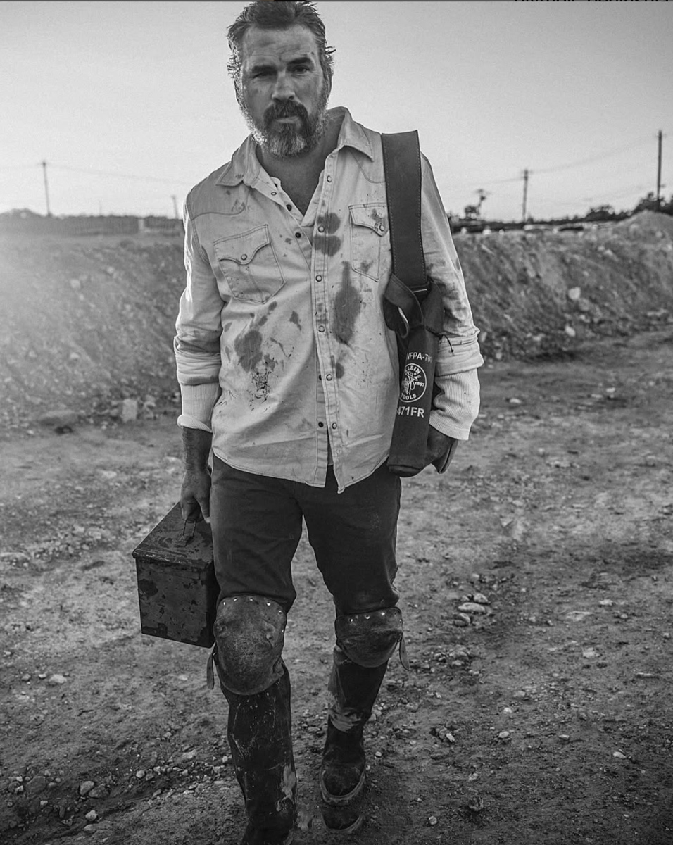 A bearded man in a dirty shirt and pants walks on a rocky, dusty path carrying a metal box and a shoulder bag. The image is black and white, and the scene looks rugged and intense.