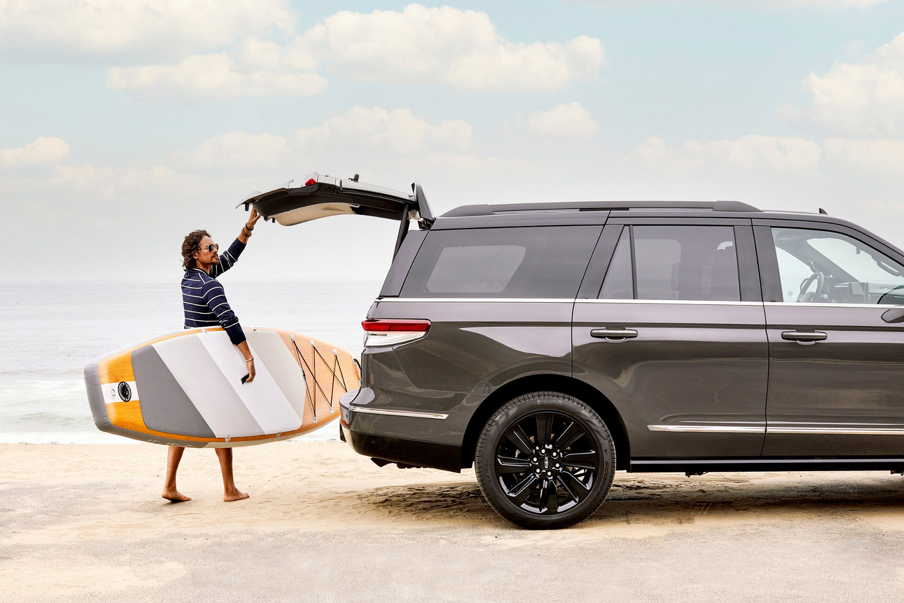 Automotive photographer Marquis Perkins captures a person holding a paddleboard beside a dark SUV with its rear hatch open on a sandy beach, the ocean and cloudy sky forming a striking backdrop.