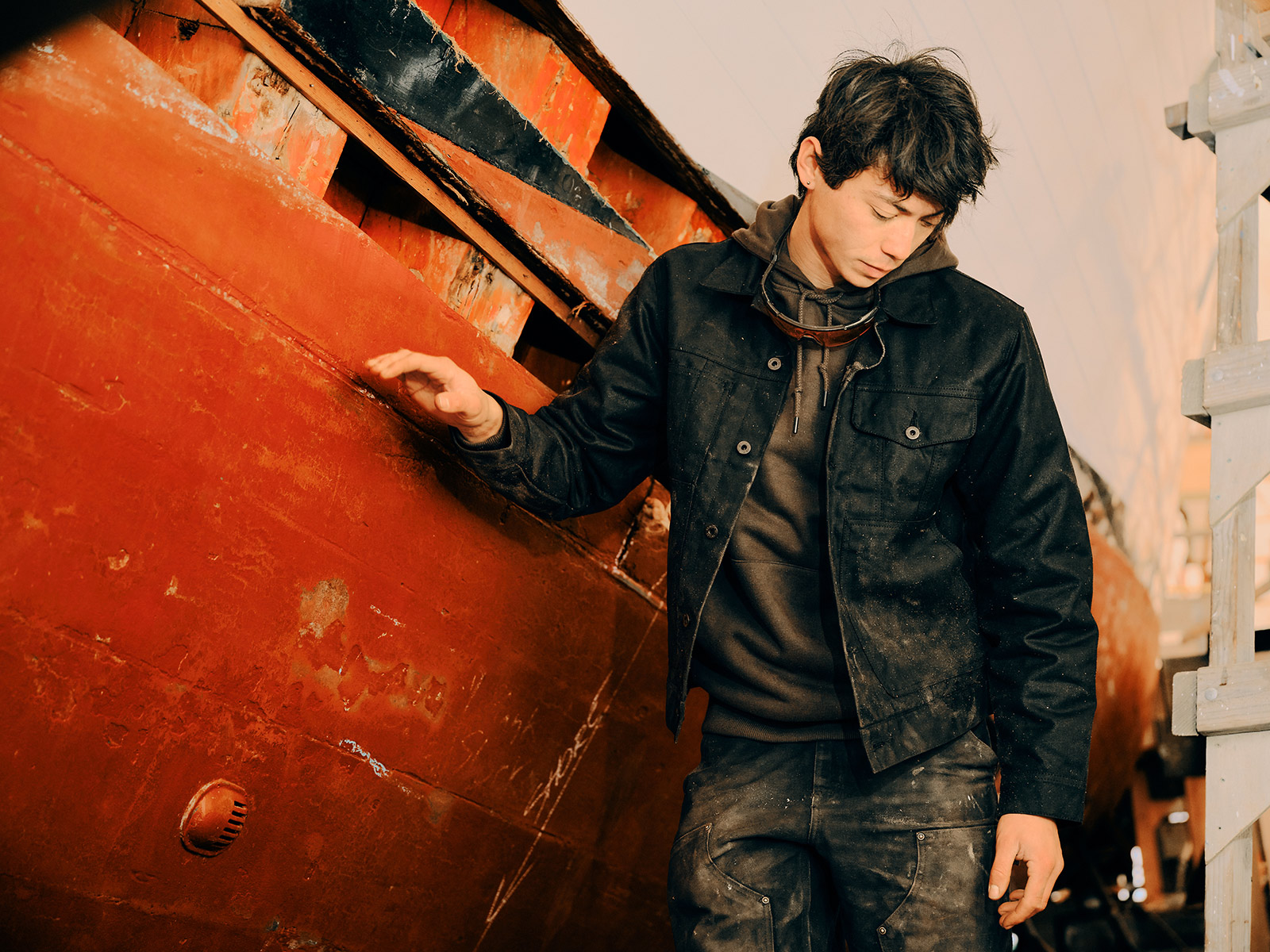 A person in work clothes stands by a weathered red boat, looking down with one hand on the hull. Captured by Portland fashion and apparel photographer Travis Gillett, the scene hints at industrial or boat repair surroundings.