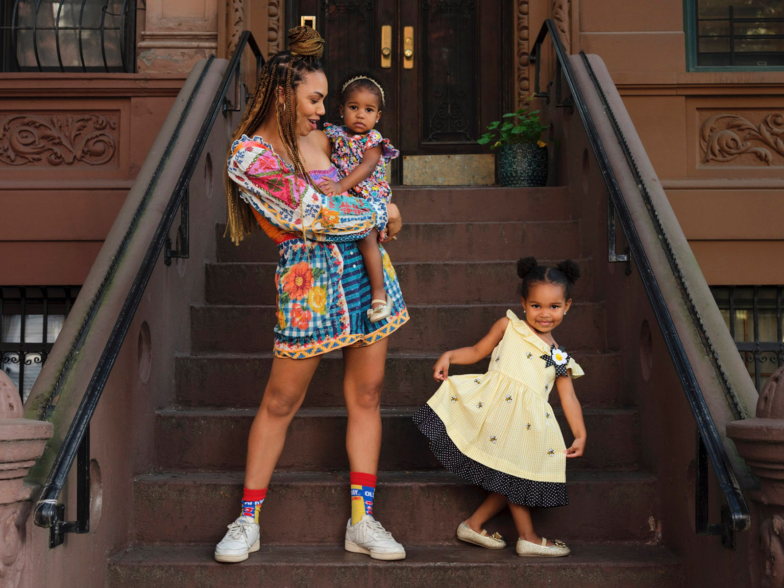 A woman in a colorful dress stands on brownstone steps, holding a baby in matching clothes. Captured by New York fashion photographer Marquis Perkins, this celebrity portrait radiates happiness and style against iconic city steps.
