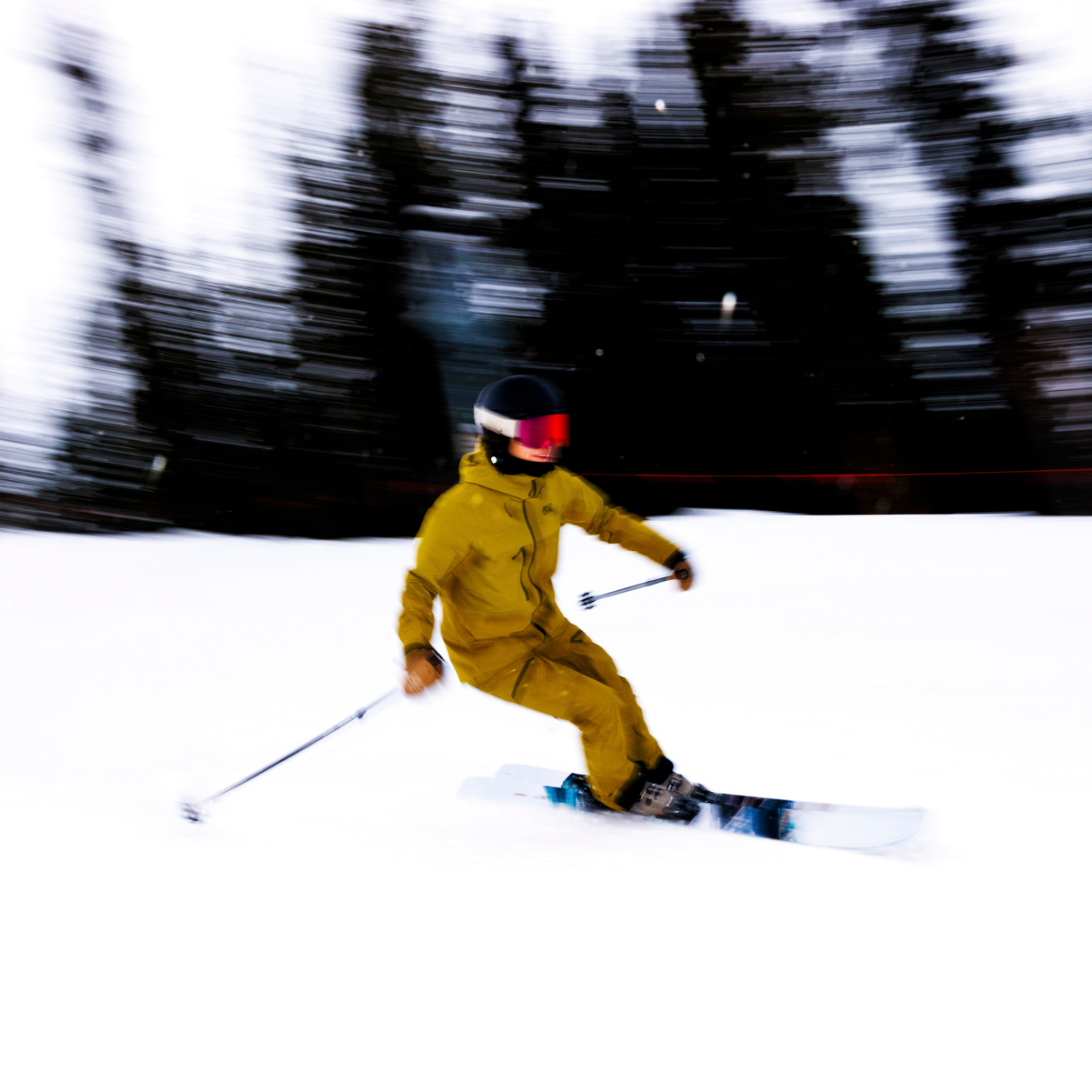A skier in a yellow suit and black helmet swiftly carves down a snowy slope, embodying the thrill of winter sports. The blurred trees in the background capture the sense of motion Sofia Jaramillo often highlights.