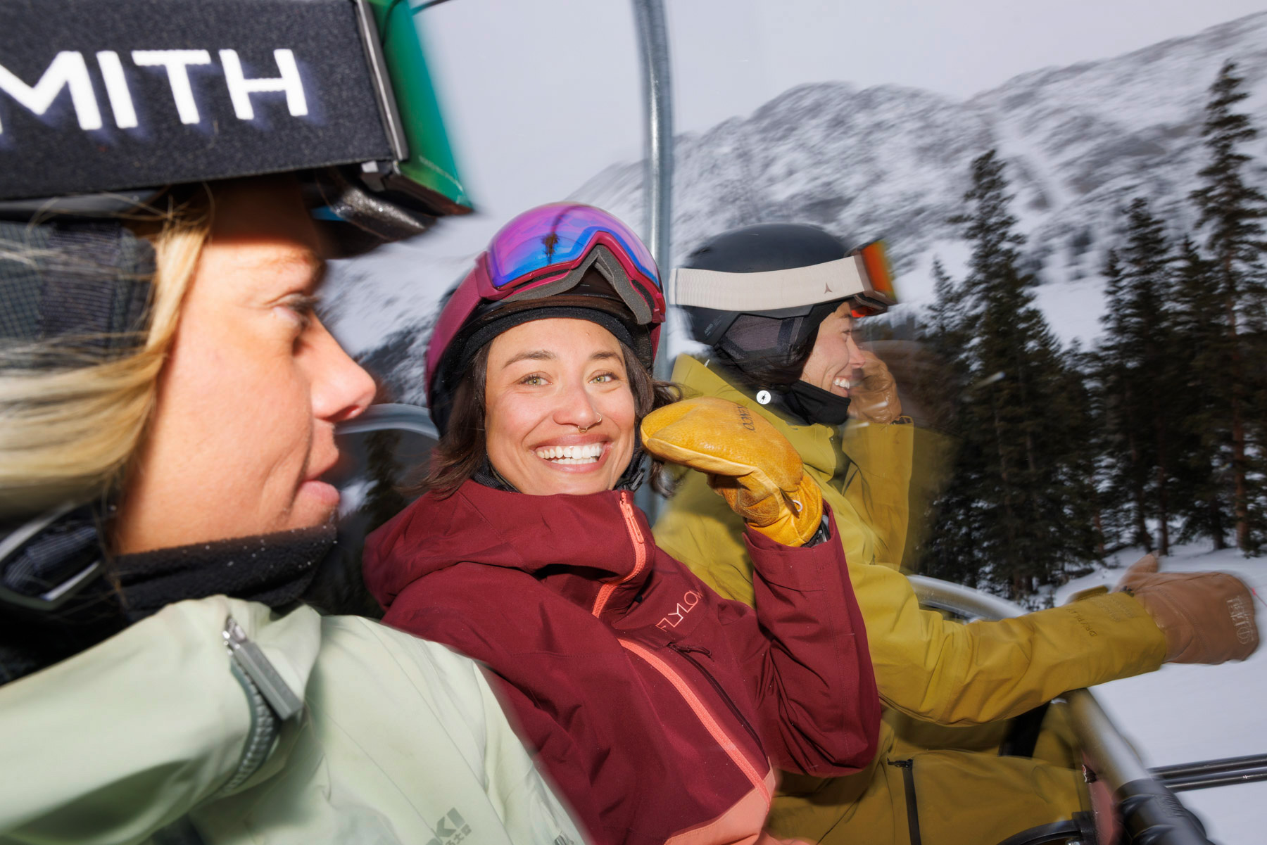Three people in winter gear ride a ski lift amid snowy mountains and trees. Sofia Jaramillo, a renowned ski photographer, captures one woman in a red jacket smiling brightly at the camera while the others look ahead.