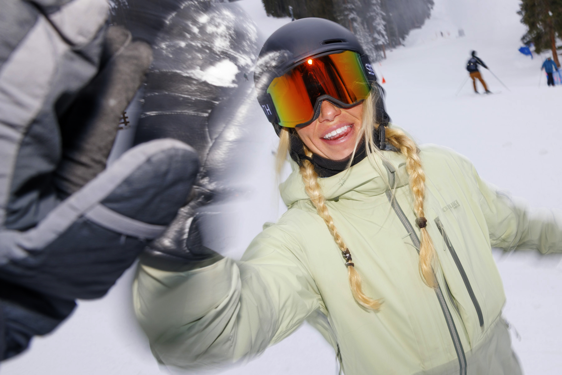 A smiling woman in a helmet and ski goggles gives a high-five on a snowy mountain, celebrating winter sports in a light green jacket with blonde braids. Other skiers and snow-covered trees are visible, perfect for a commercial photographer’s portfolio.