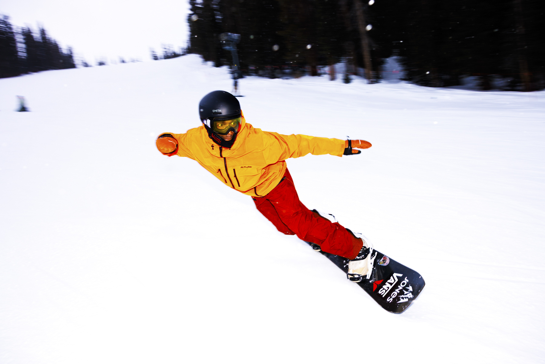 A snowboarder in a yellow jacket and red pants glides down a snowy Idaho slope, leaning sharply with one arm extended, surrounded by trees in the background—a perfect scene for winter sports enthusiasts.
