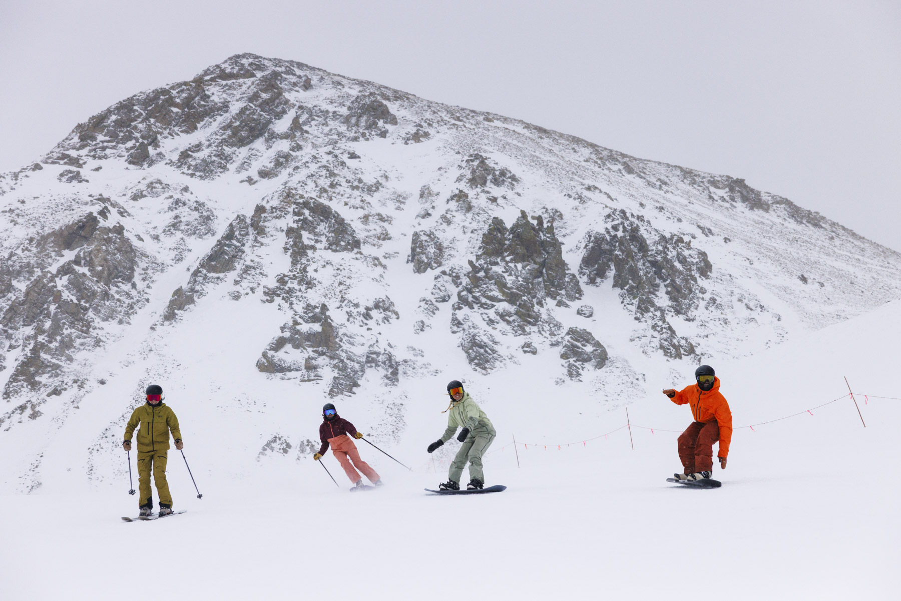Four people, dressed in colorful winter gear, ski and snowboard down a snowy mountain slope beneath a rocky, snow-covered peak on a cloudy day, captured by commercial photographer Sofia Jaramillo.