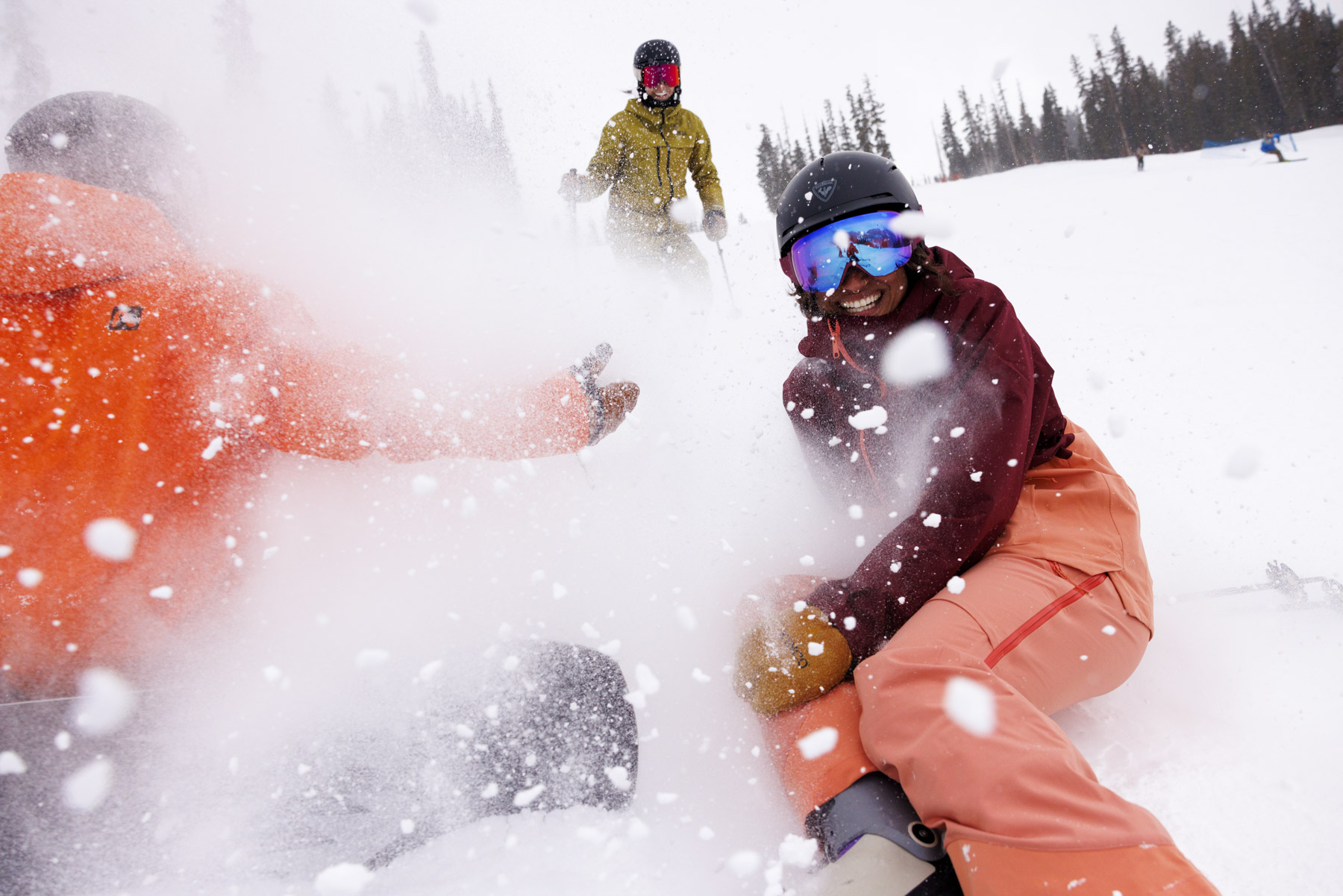 Three people in winter gear enjoy the snow on an Idaho ski slope, with fresh powder flying in the air. One person in orange and pink smiles at the camera, captured by a commercial photographer, while others stand and sit among snowy trees.