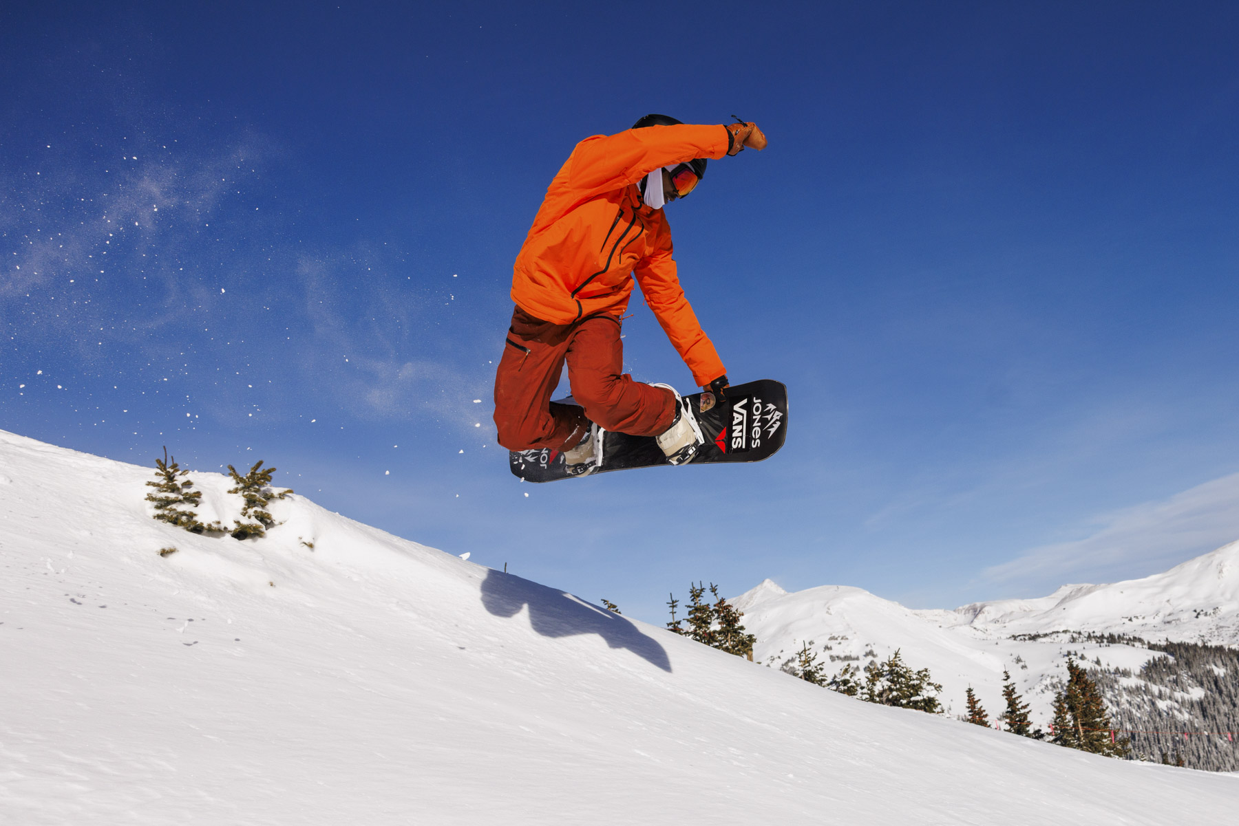 A snowboarder wearing an orange jacket and red pants jumps into the air over a snowy Idaho slope, with clear blue sky and snow-covered mountains in the background, capturing the thrill of winter sports.