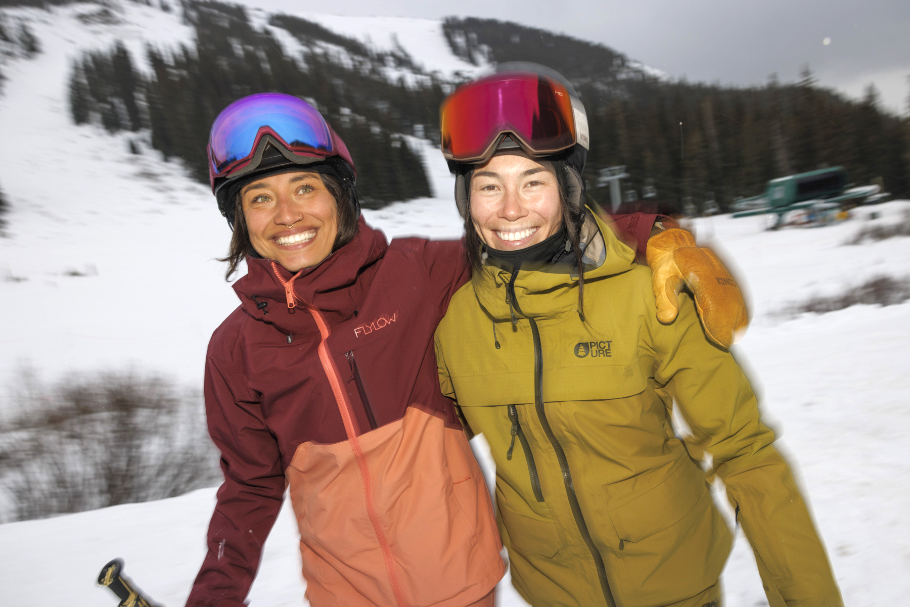Two smiling people wearing ski jackets, gloves, and helmets stand together with arms around each other in a snowy Idaho mountain landscape, with ski slopes and trees in the background.