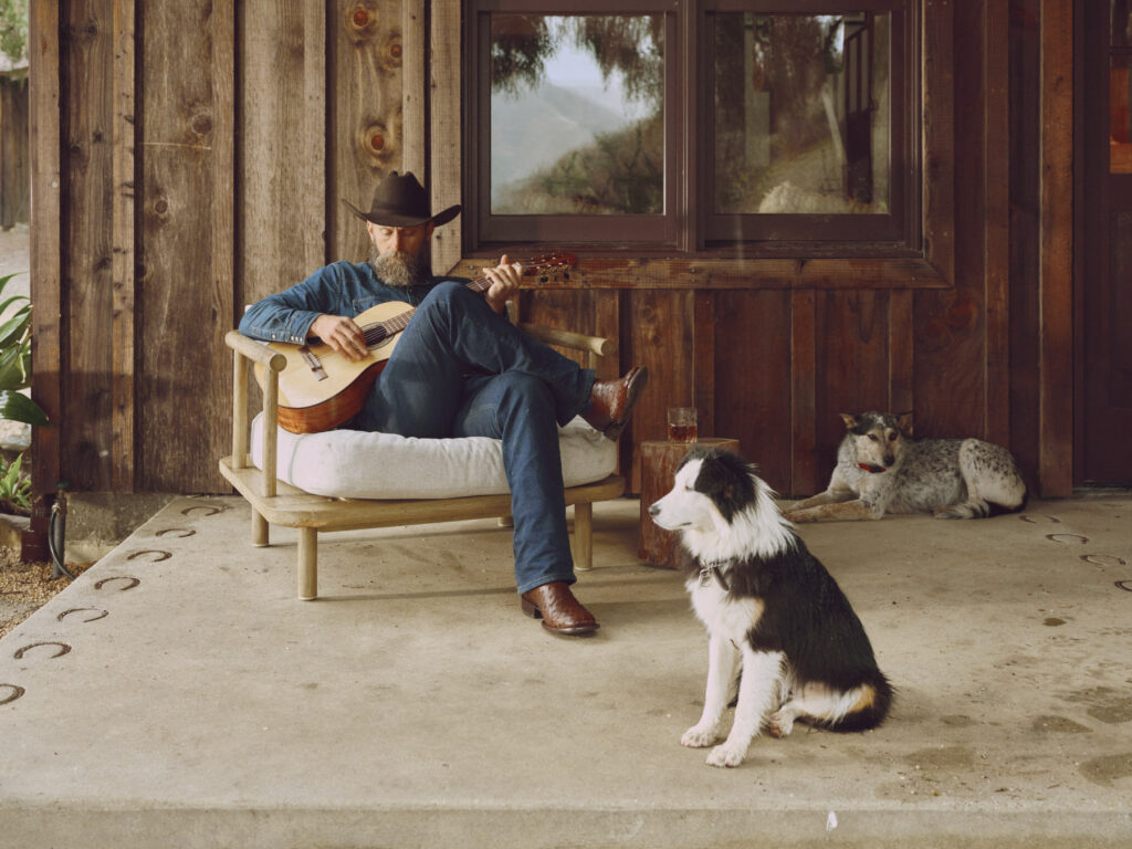 A man in a cowboy hat, wearing Tecovas boots, plays guitar on a cushioned chair on a rustic porch. A black and white dog sits nearby, while another rests in the corner by the wooden wall.