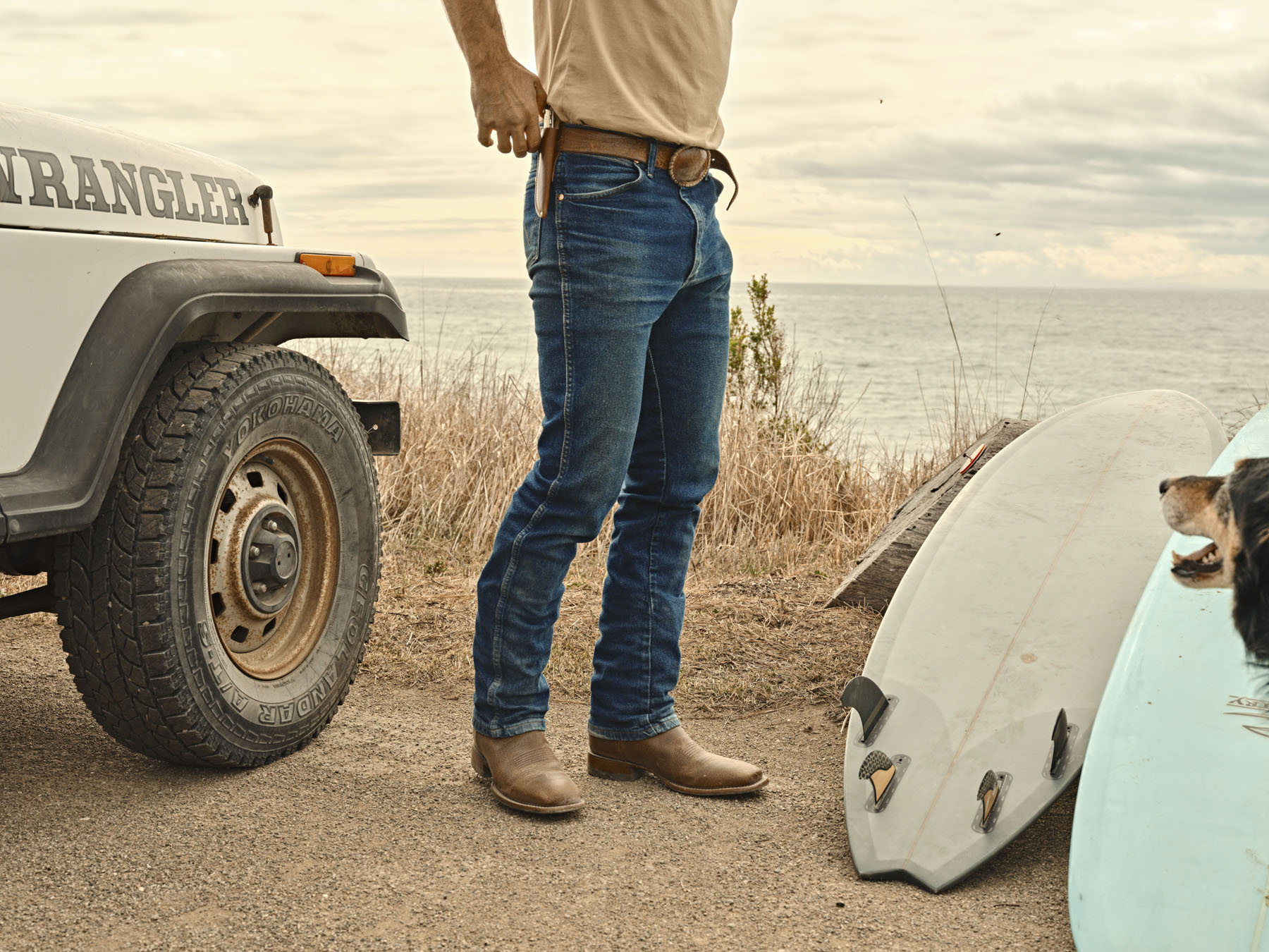 A person in workwear jeans and boots stands by a white Jeep Wrangler near the ocean, a knife in their belt. A surfboard rests on the ground as a black dog looks up—captured through the lens of Los Angeles photographer Travis Gillett.