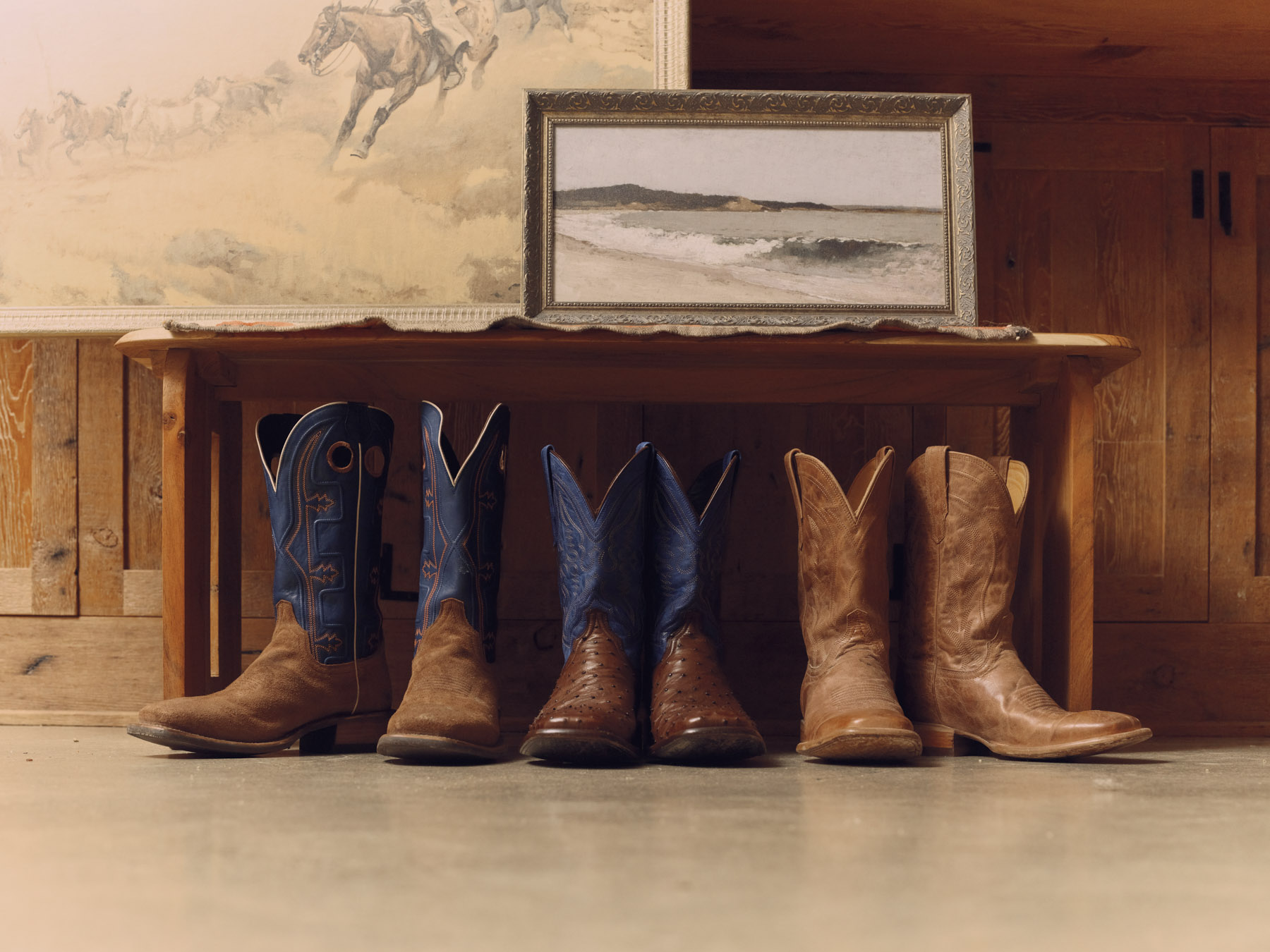 Four pairs of Tecovas cowboy boots are lined up on the floor in front of a wooden bench, with two framed landscape paintings above them. Captured by Los Angeles photographer Travis Gillett, the setting exudes rustic, cozy charm.