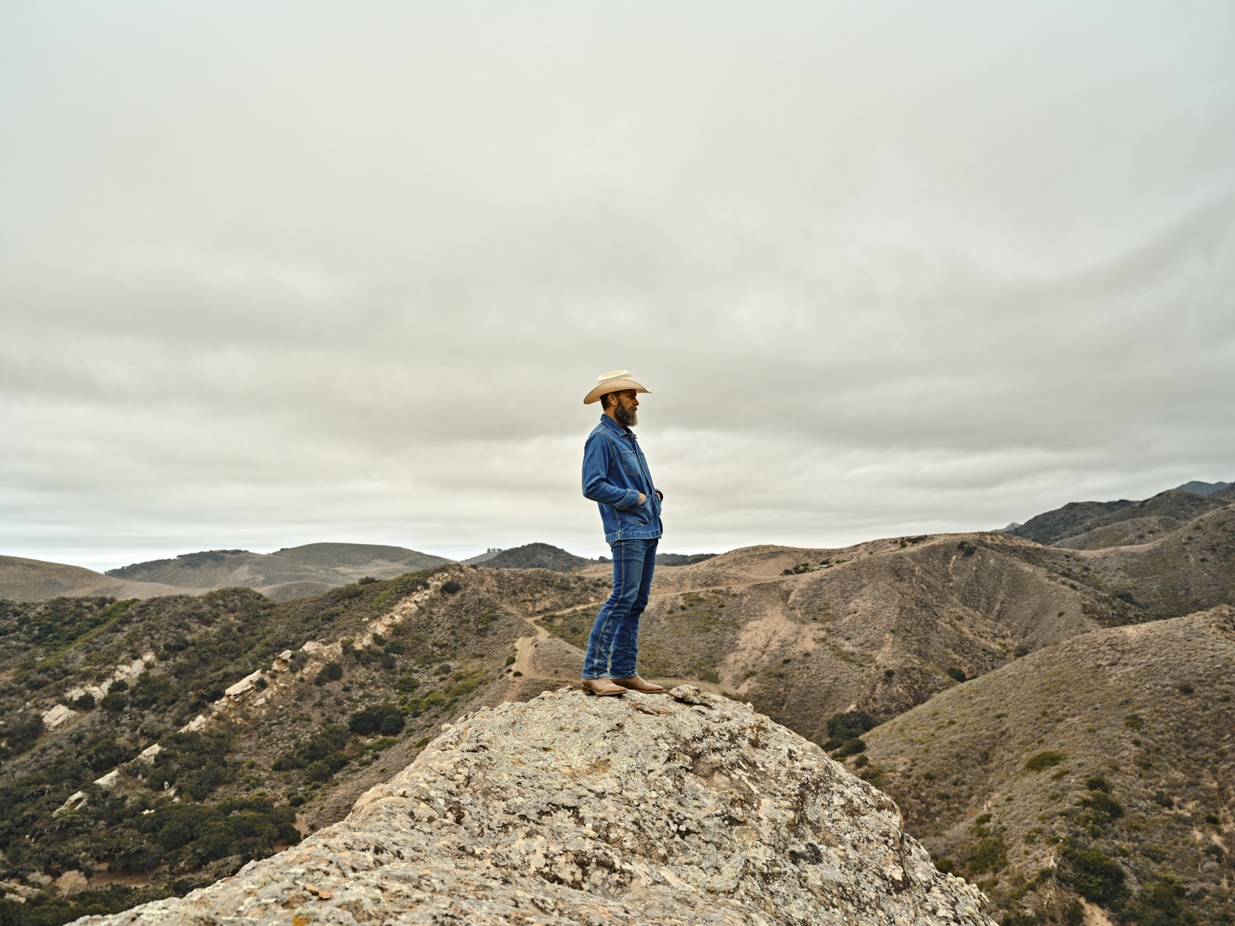 A person wearing a cowboy hat, blue jacket, and jeans stands on a rocky outcrop, overlooking rolling hills beneath a cloudy sky—captured by Los Angeles photographer Travis Gillett.