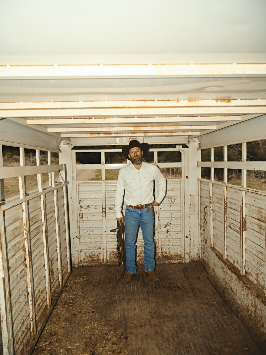 A man wearing a black cowboy hat, white shirt, blue jeans, and Tecovas boots stands inside a weathered, empty livestock trailer, holding a rifle—captured with the keen eye of a Los Angeles fashion photographer.