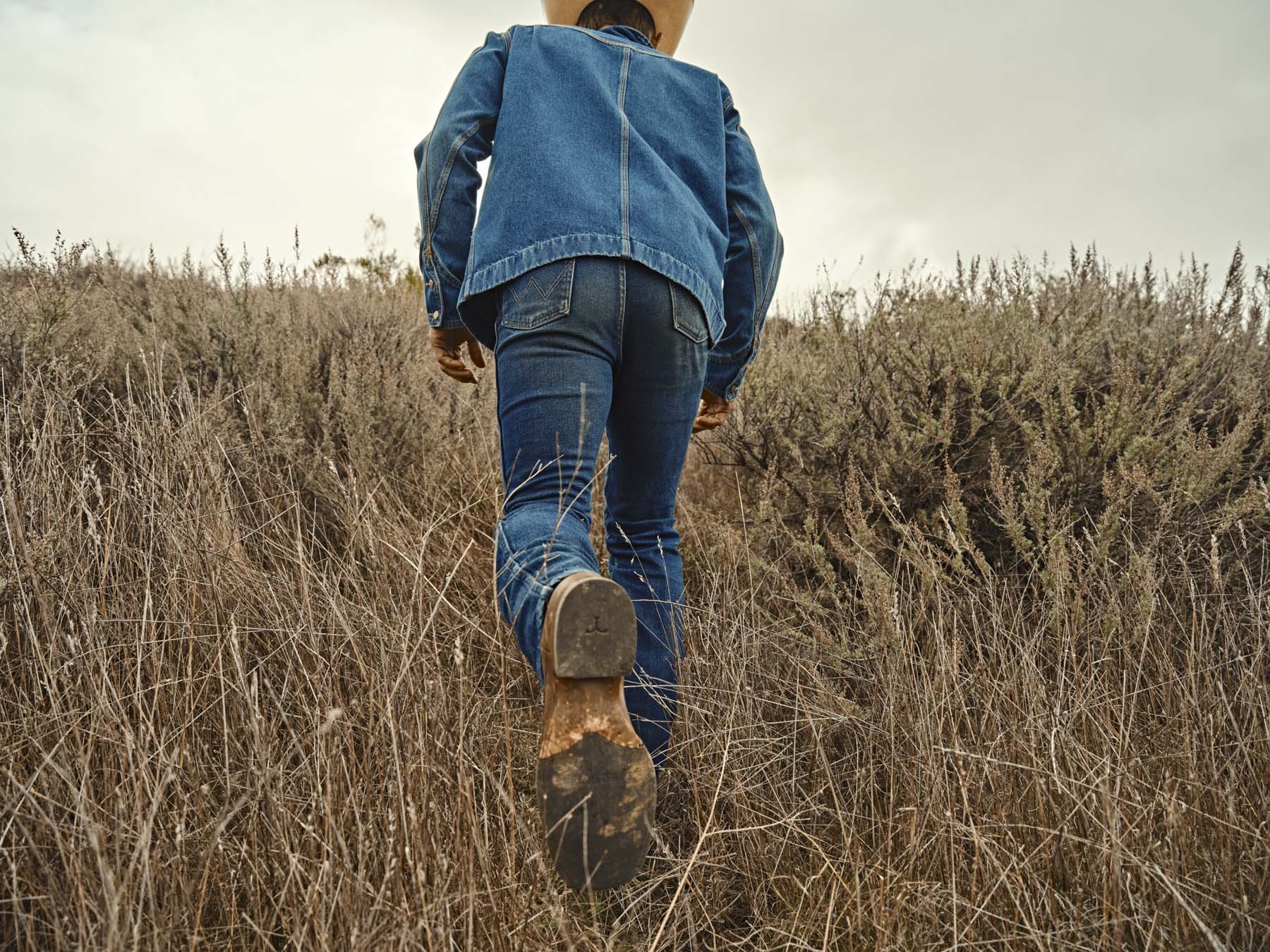 Photographed by Los Angeles photographer Travis Gillett, a person in workwear—jeans, denim jacket, and cowboy hat—walks uphill through dry grass. Captured from behind at ground level, the rustic landscape and overcast sky set a natural mood.