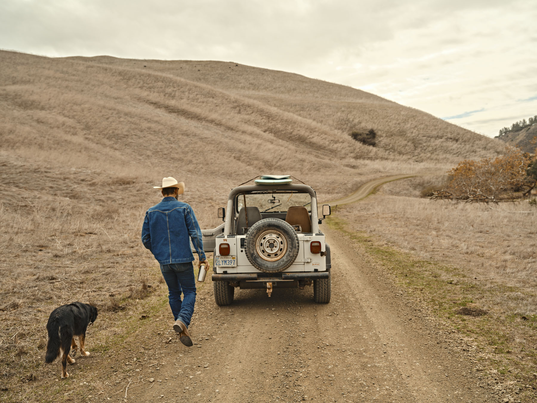 A person in a denim jacket and hat, channeling classic Tecovas style, walks with a black dog on a dirt road beside a white off-road vehicle in a dry, grassy landscape with rolling hills beneath a cloudy sky.