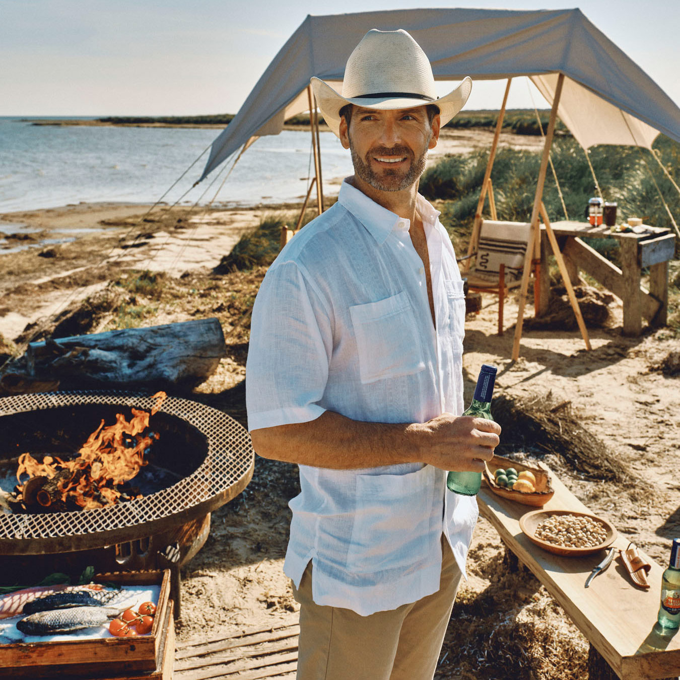 A man in a white shirt and cowboy hat stands by a fire pit near a beach, holding a drink. Captured by lifestyle photographer Travis Gillett, he smiles with food and drinks on a nearby table and a tent set up in the background.