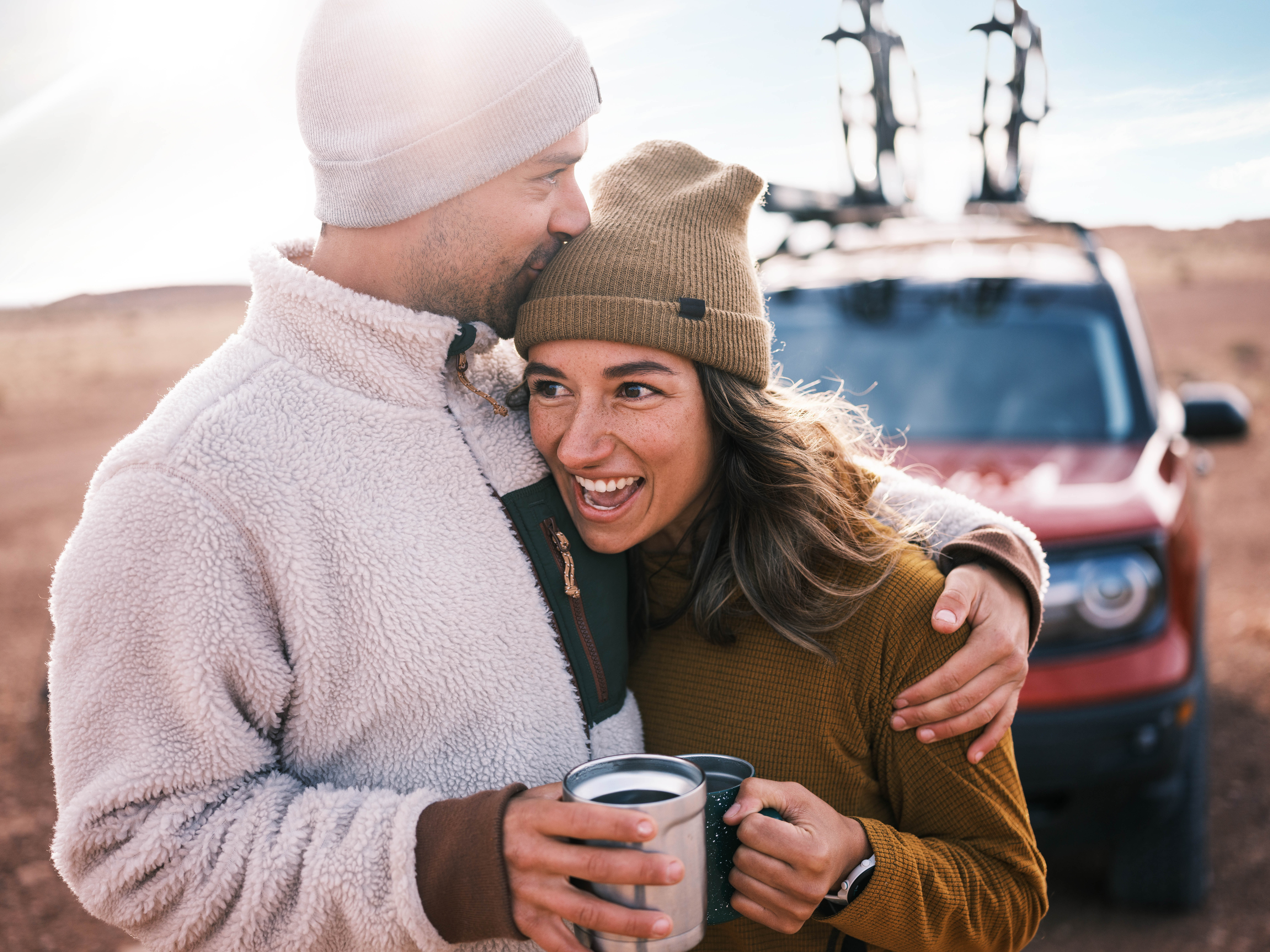 A smiling couple wearing warm clothing stands outdoors, embracing and holding metal mugs. Behind them is a red Bronco SUV with bikes mounted on the roof. The sun shines brightly in the background, capturing a classic Sun Valley adventure moment for any photographer.