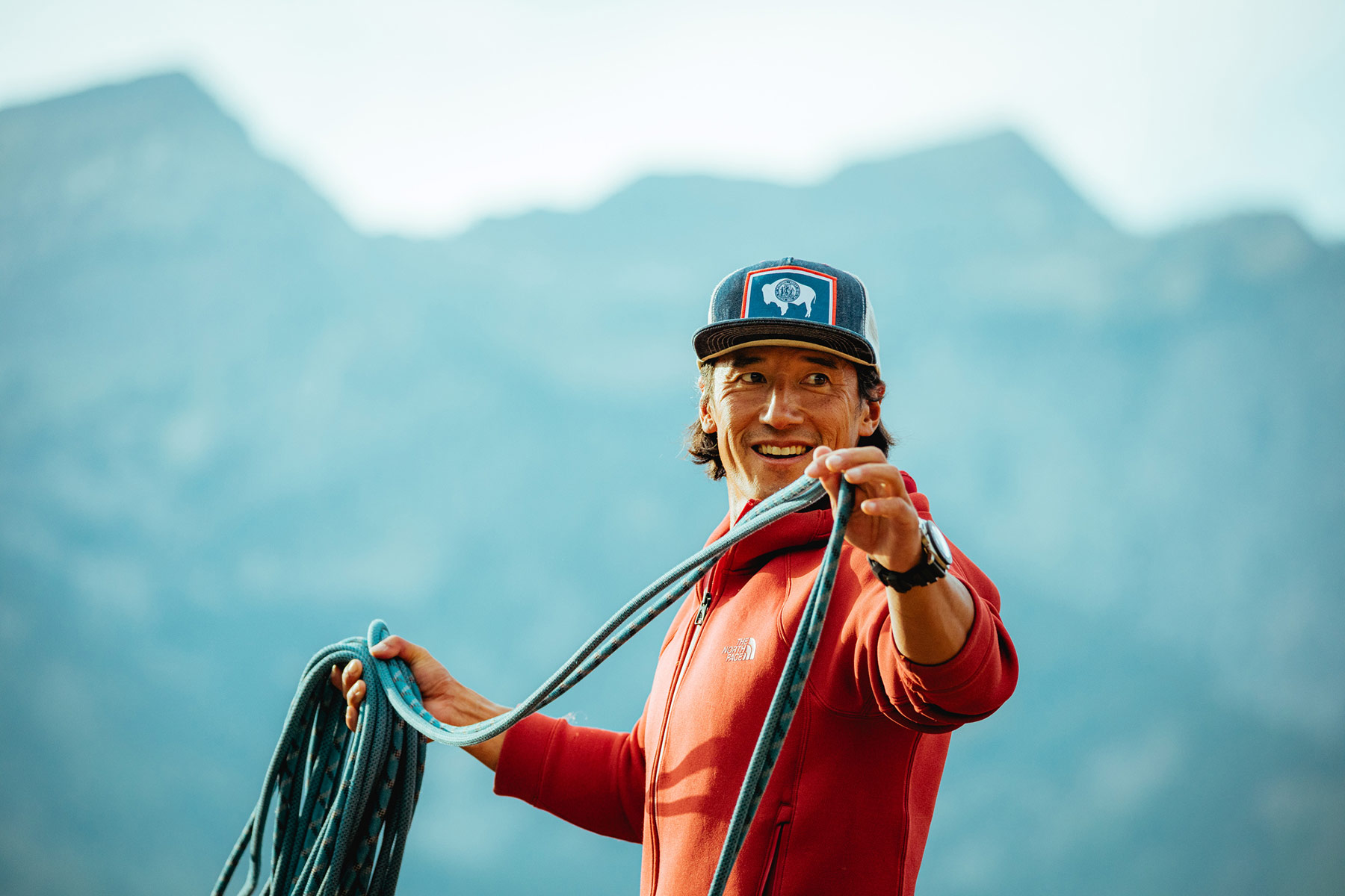 A person in a red jacket and cap stands outdoors in Sun Valley, smiling while holding and coiling a climbing rope. The background is out of focus, emphasizing the photographer’s expression and activity.