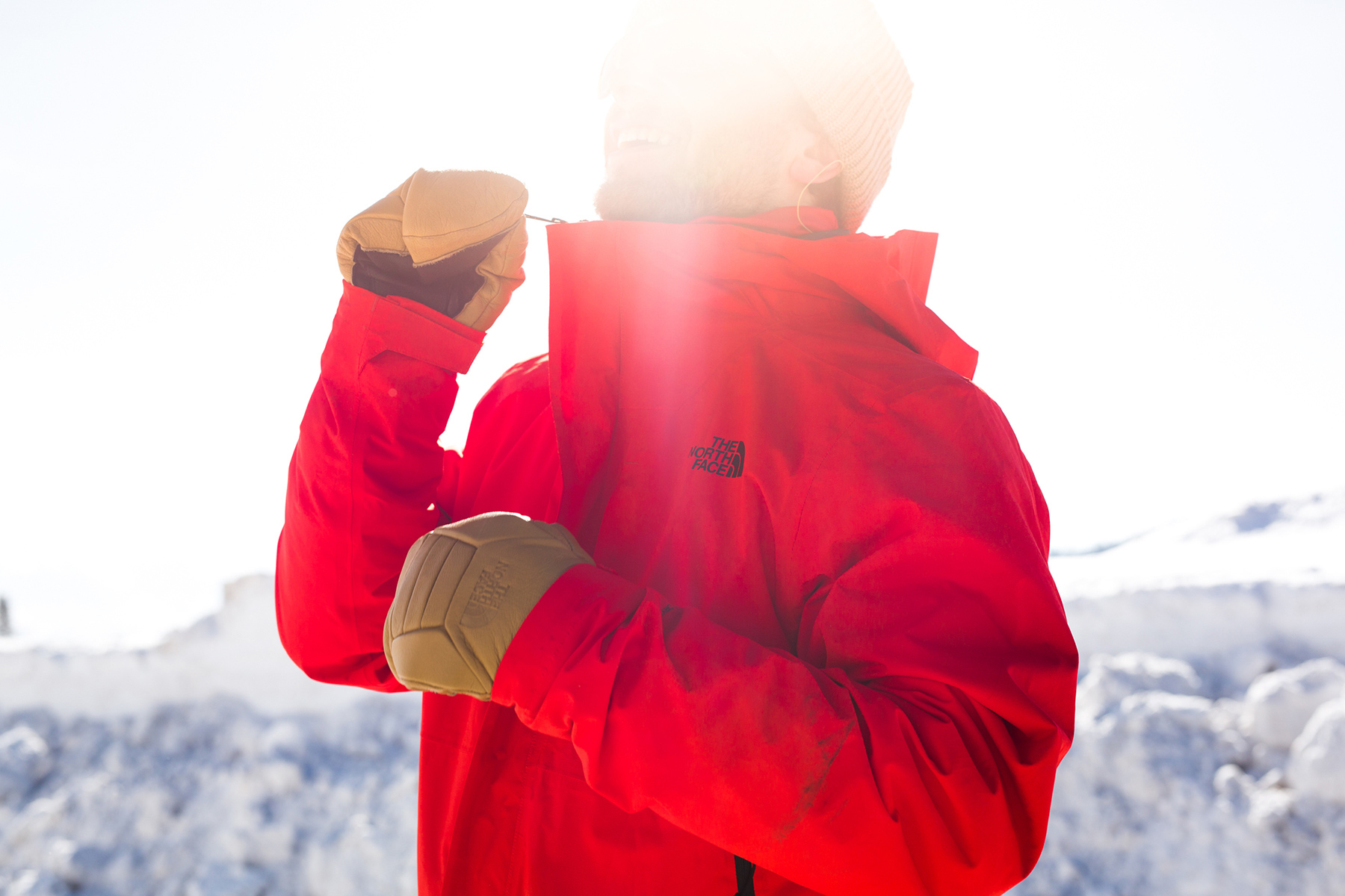 A person in a red North Face jacket and tan gloves zips up their coat in a snowy, sunlit outdoor setting. The bright sunlight partially obscures their face, with snow-covered trees behind, as captured by apparel photographer Sofia Jaramillo.