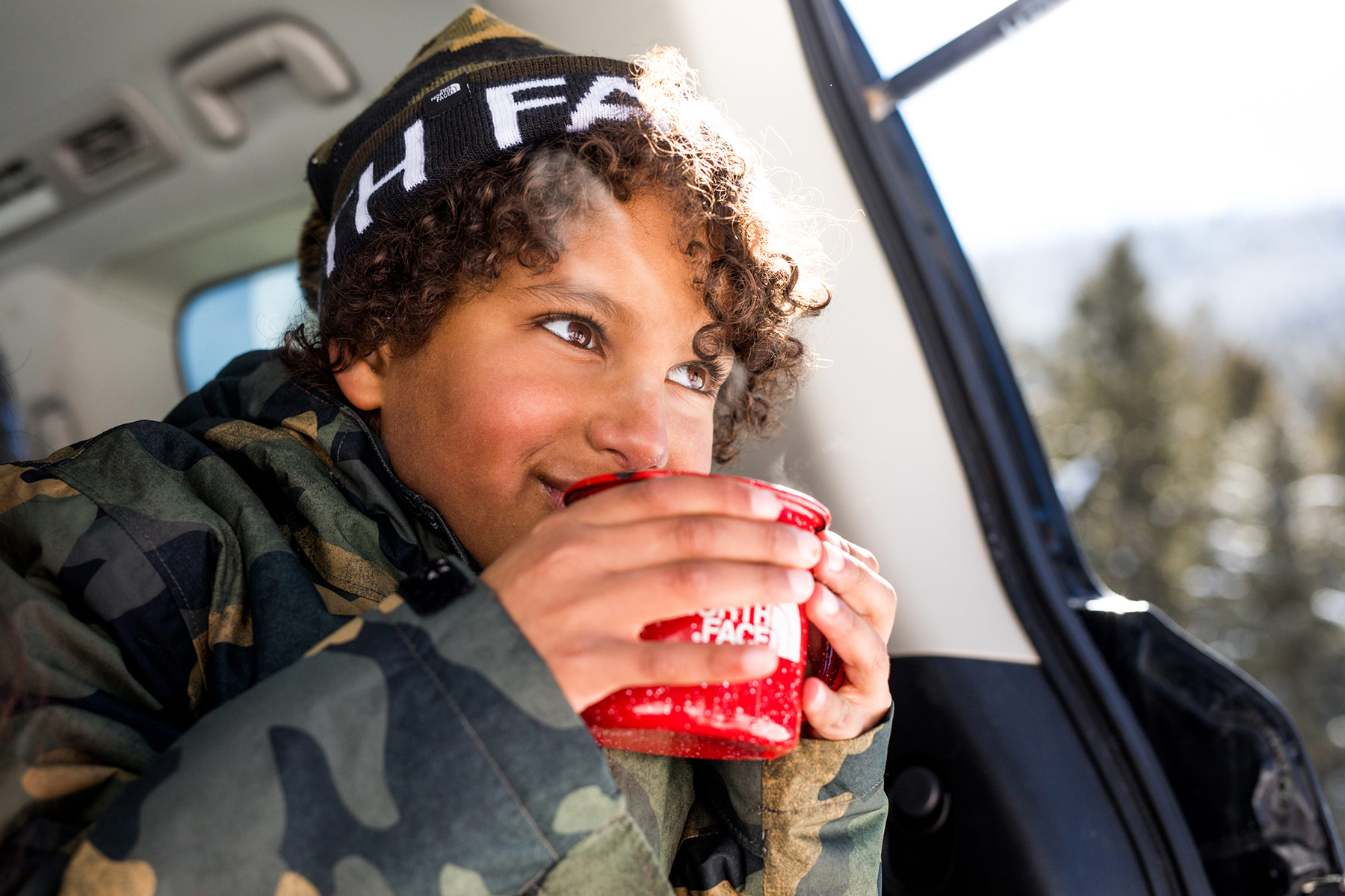 A child wearing a camouflage jacket and a black beanie sips from a red mug inside a vehicle, with snow-covered Idaho trees visible outside the window.