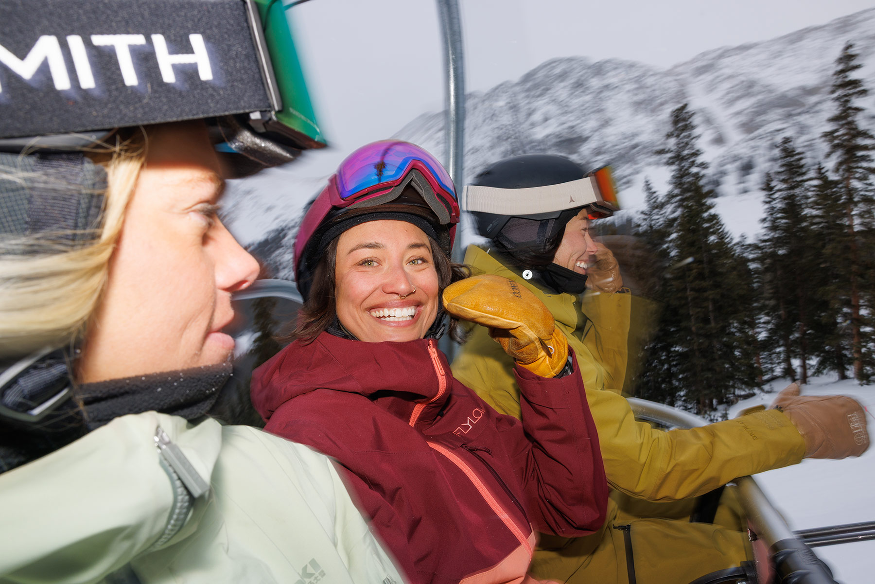 Three people in winter jackets and helmets ride a ski lift with snowy Idaho mountains and pine trees in the background. The person in the middle smiles excitedly at the camera, wearing ski goggles and brown mittens.