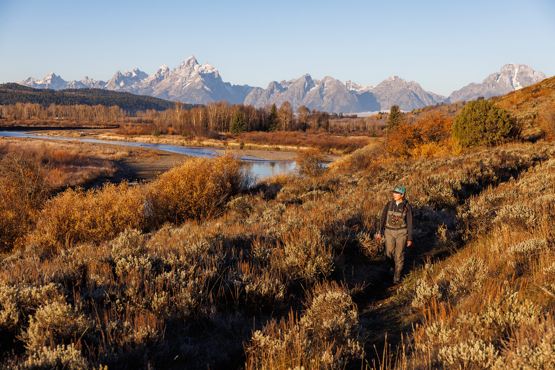 A person in outdoor apparel hikes along a sunlit trail through autumn shrubs, with a river and Idahos snow-capped mountain range visible in the background under a clear blue sky.