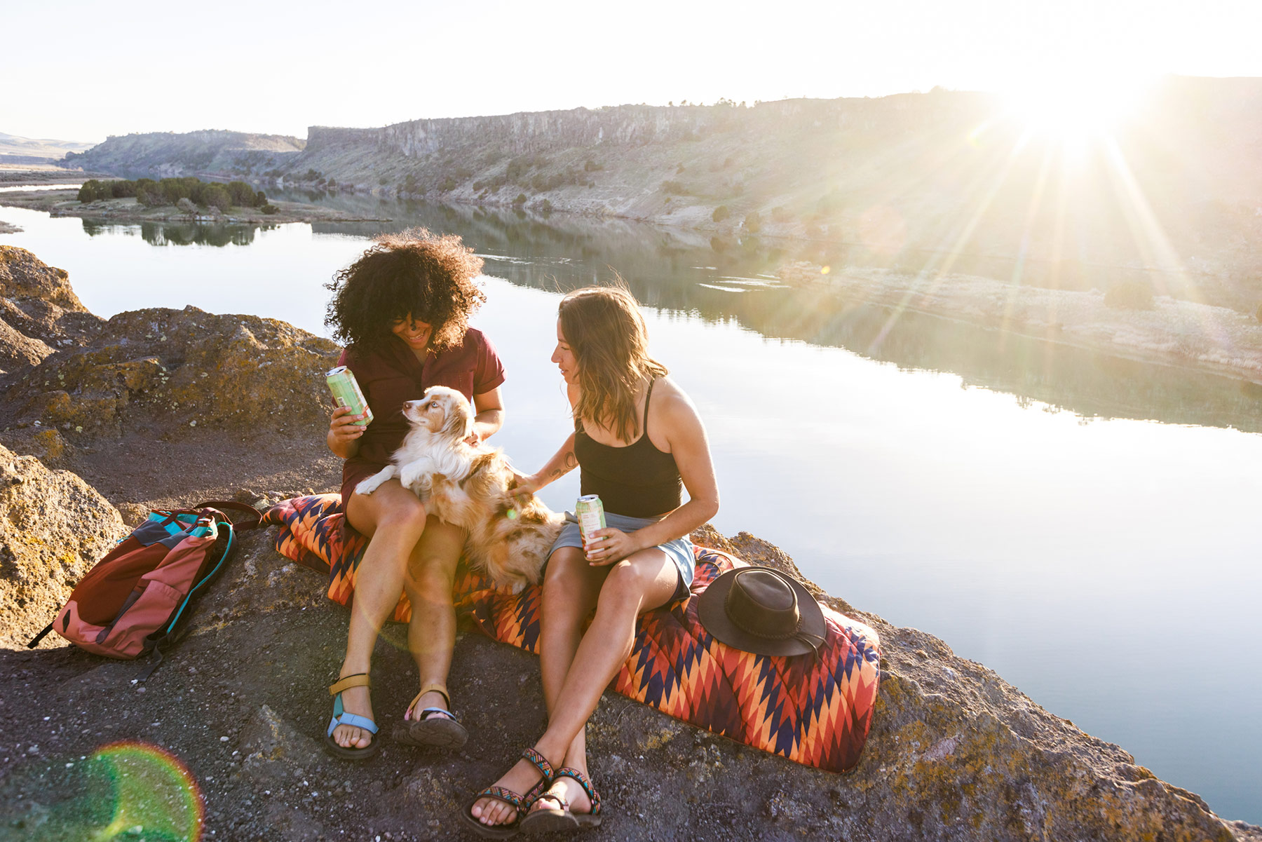 Two women sit on a colorful blanket by a river at sunset, smiling and holding drinks, with a dog on their laps. Unrivaled outdoor adventure photographer vibes are captured as a backpack and hat rest nearby on the rocky ground.