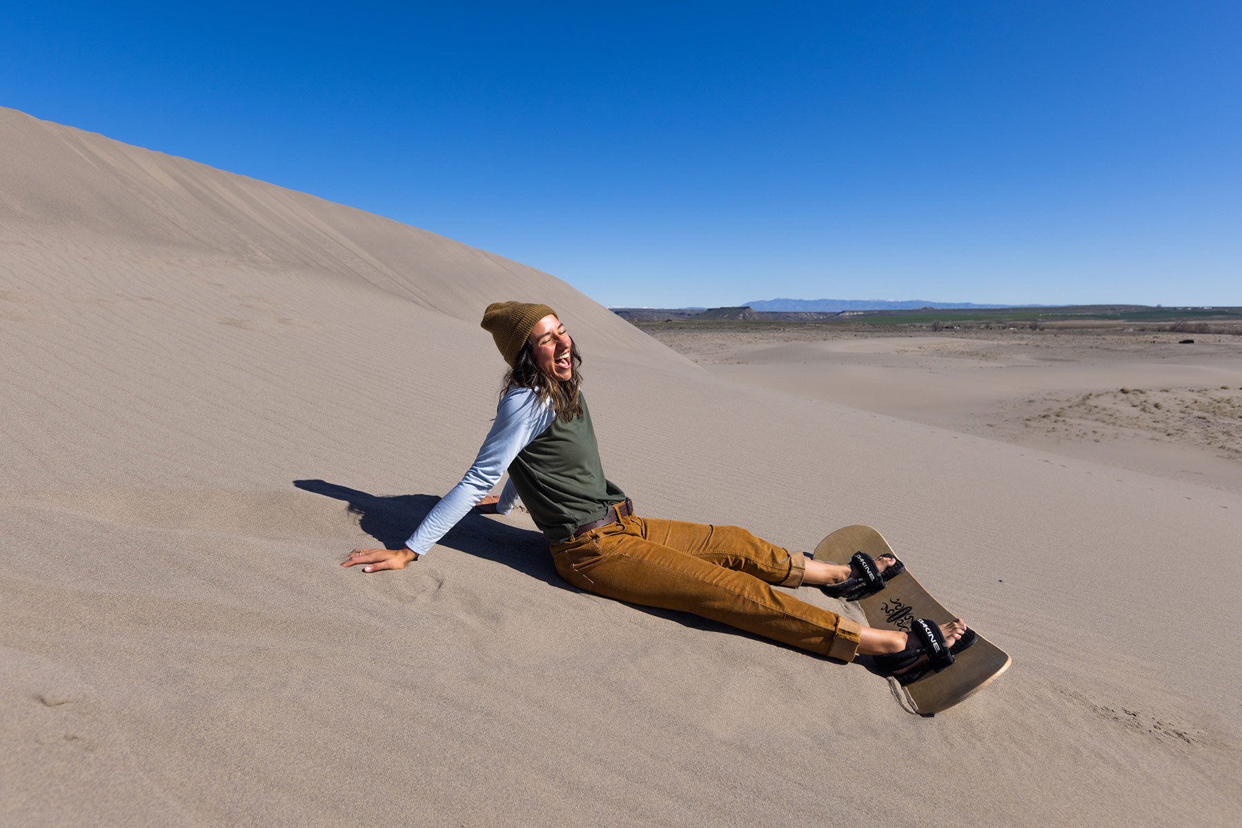 A person wearing outdoor apparel—a beanie, long-sleeve shirt, and pants—sits on a sand dune with a sandboard, smiling under a clear blue sky in a desert landscape. Photo by Sofia Jaramillo.