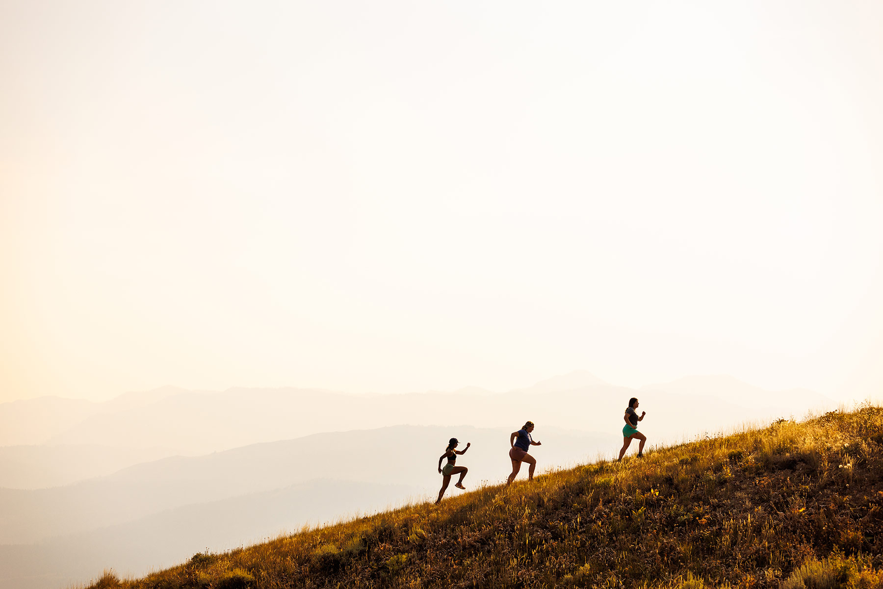 Three people in outdoor apparel are trail running uphill on a grassy slope during sunset or sunrise, with hazy mountains and a bright sky in the background.
