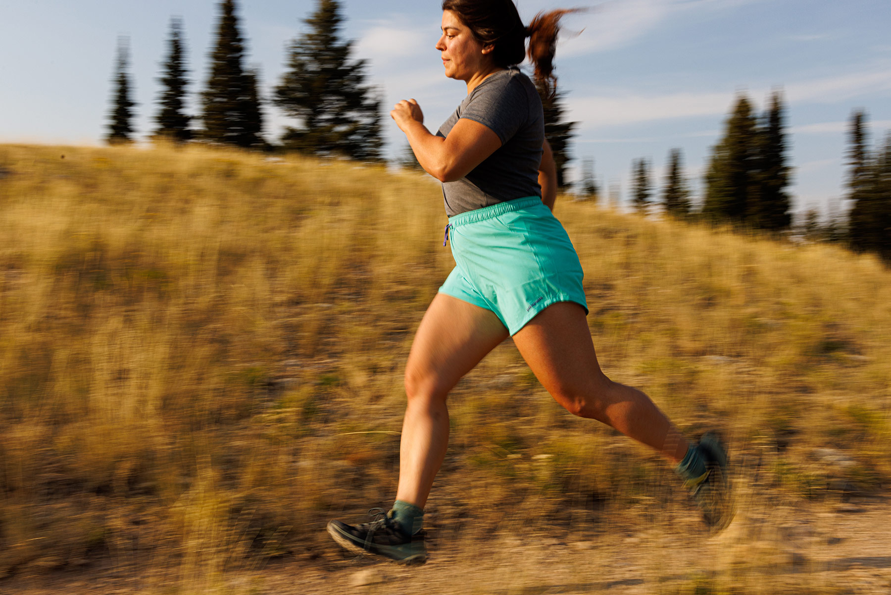A woman in a gray t-shirt and turquoise shorts runs energetically on a dirt trail in a grassy, sunlit Idaho landscape, captured by outdoor adventure photographer Sofia Jaramillo with trees in the background.